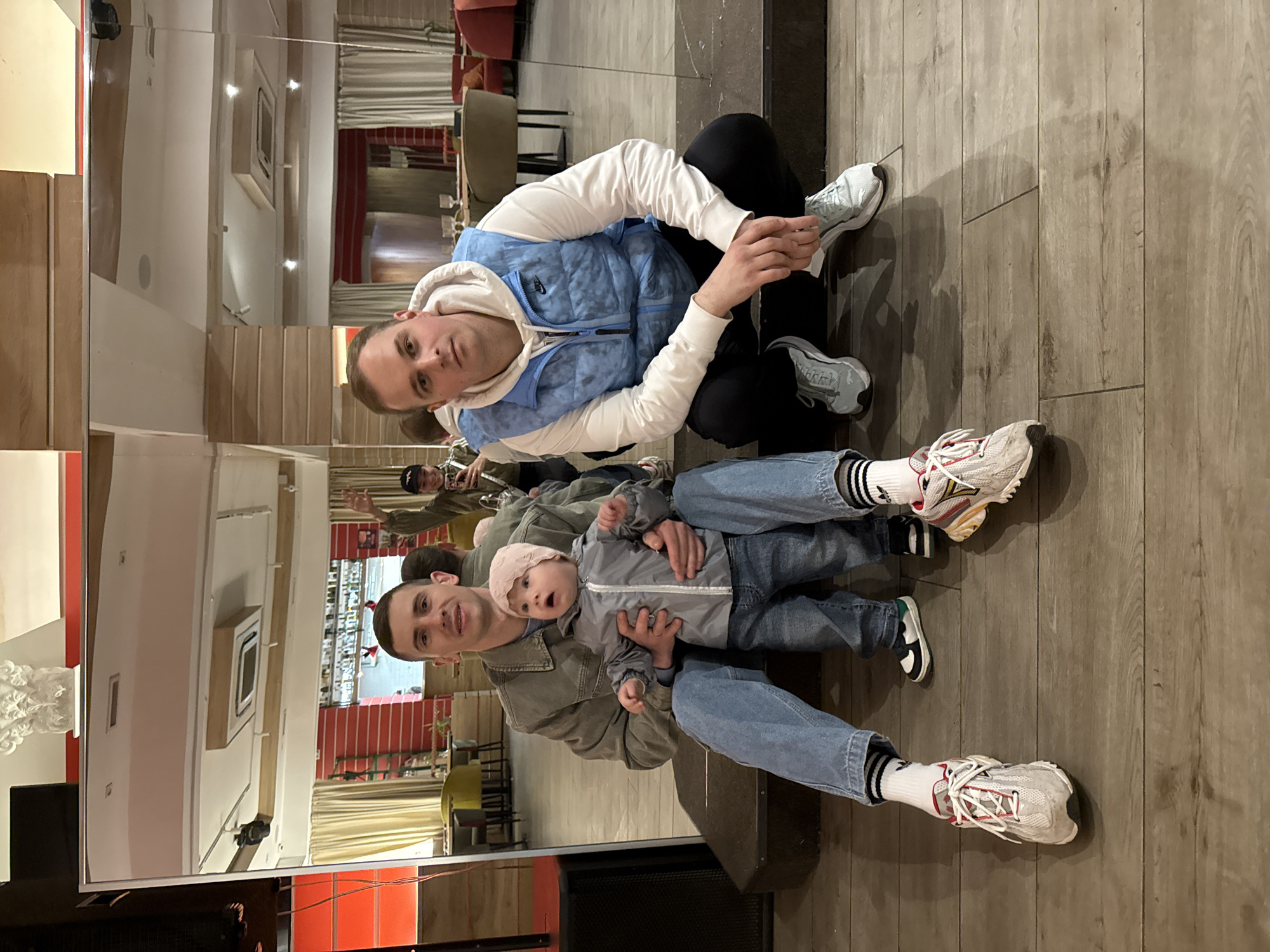 A man and a baby are reflected in a mirror. The man, wearing jeans and a jacket, is holding the baby. His reflection is seen in the mirror. In the reflection, the man is wearing black pants and a white hooded shirt with a light blue vest. The reflection also shows the interior of a restaurant. The restaurant appears to be in Moscow, Russia based on the reflection of red-themed interior design in the mirror, which is typical for Moscow restaurants.