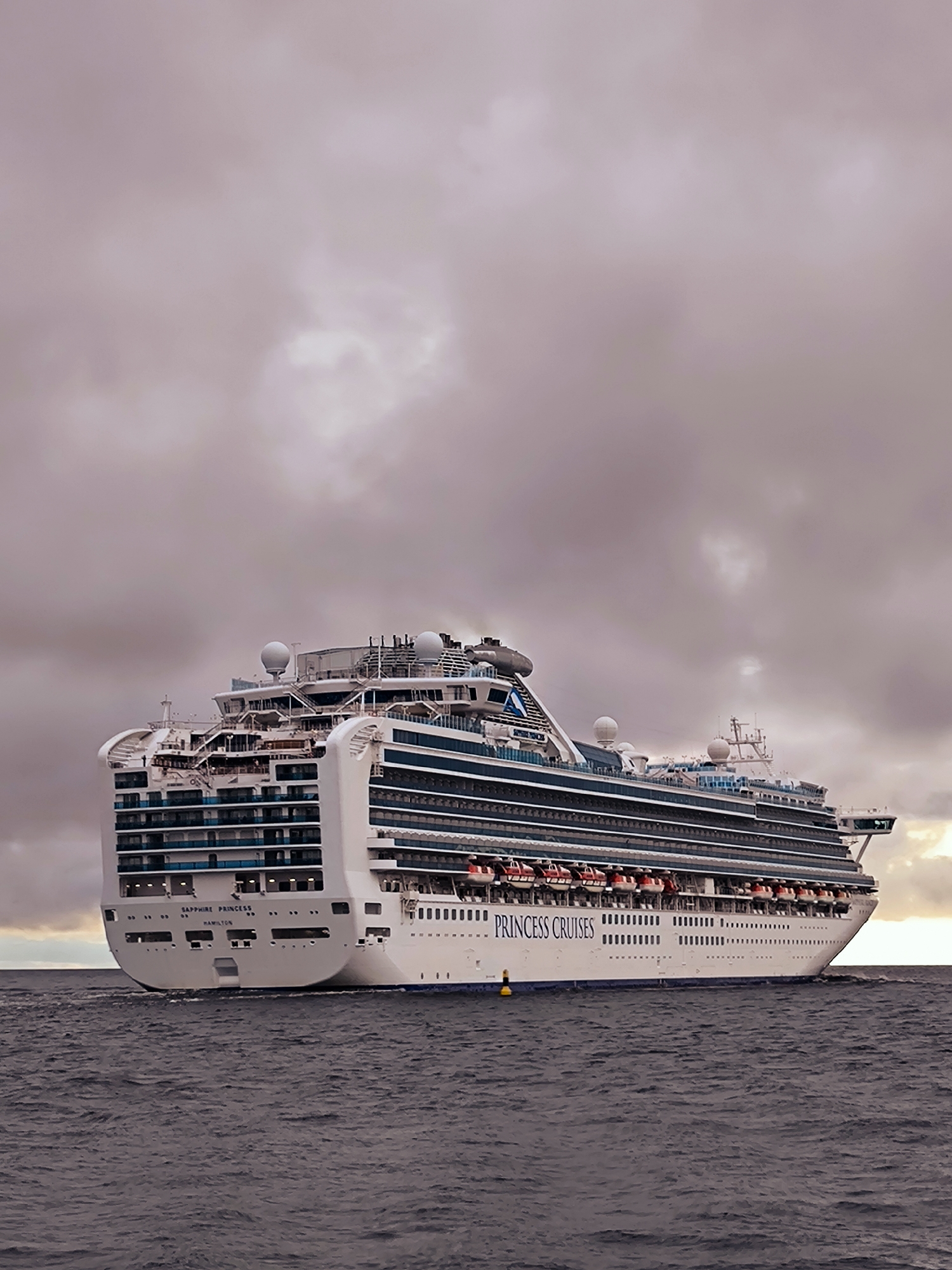 A large, white cruise ship named "SAPPHIRE PRINCESS" and "PRINCESS CRUISES" is sailing on a dark, choppy body of water under a cloudy, overcast sky. The ship is viewed from its stern and port side, revealing multiple decks with balconies, lifeboats, and various antennas and structures on its upper levels. The ship's name and homeport, "HAMILTON," are visible on the stern. A yellow buoy is floating in the water near the ship. The overall scene is one of a maritime vessel in open water, with no visible land features or cityscapes. The location in Riga, Latvia, is not depicted in this image.