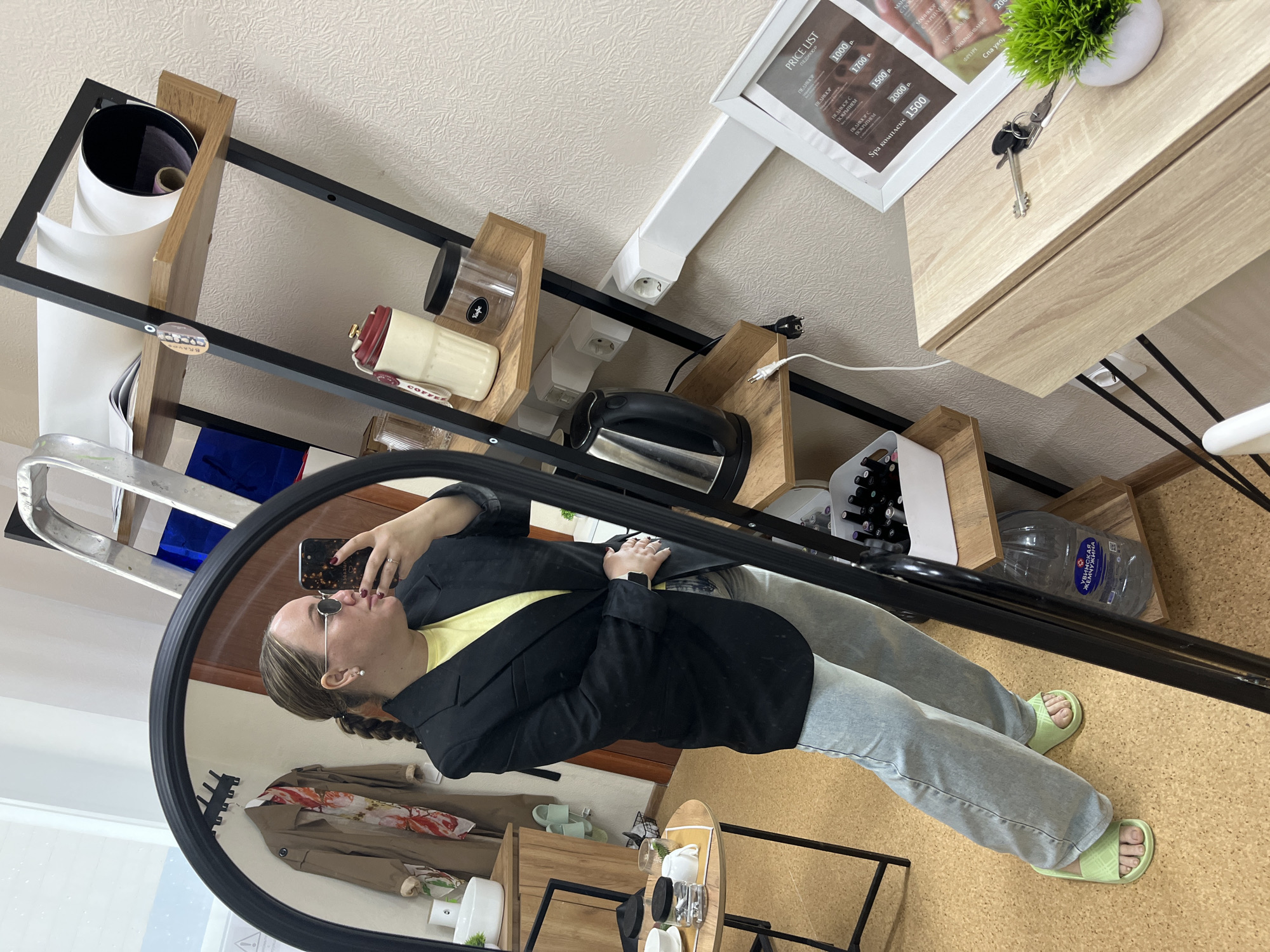 The image depicts a person taking a mirror selfie in what appears to be an indoor setting, possibly an apartment or a shop. The person is wearing a black blazer over a yellow shirt and grey jeans, with green sandals on their feet. They are holding a smartphone in their hand, taking the photo in front of a round mirror with a black frame.
In the background, a shelving unit is visible, holding various items such as rolls of paper, jars, a kettle, bottles, and other miscellaneous objects. A price list is mounted on the wall, and there is a small decorative plant on a nearby surface. The flooring is cork.
The location, as indicated by the OCR text, appears to be the city of Chaikovsky, Russia, based on Cyrillic text mentioning "Жувинская" and "Кофе."