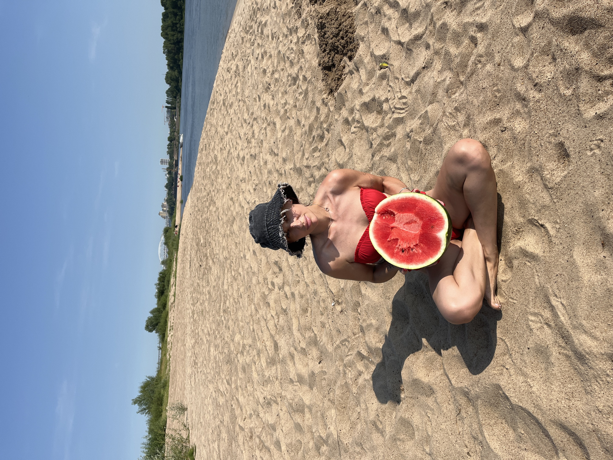 A woman wearing a red bikini and a black bucket hat sits cross-legged on a sandy beach, holding a large slice of watermelon. The beach is next to a wide river, with a city skyline visible in the distance across the water. The sky is clear and blue. The scene appears to be in the city of Nizhny Novgorod, Russia, given the presence of the river and the urban landscape across it, likely on the Volga River.