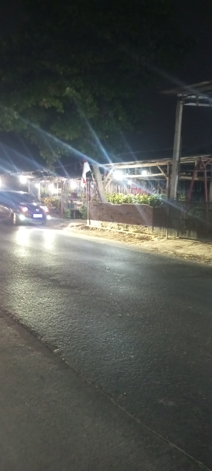 The image is a nighttime, low-light shot of a street scene in Setu, Indonesia. A dark-colored car is parked on the left side of the asphalt road, its headlights illuminated. In the background, a structure with a corrugated metal roof supported by wooden posts is visible. This structure appears to be a covered market or stall, with bright lights illuminating the interior. Rows of what appear to be bananas are displayed on a brick ledge in front of the structure. A flag, predominantly white with a red stripe on top, is mounted on a pole in front of the building. Trees with dark foliage are present in the upper portion of the image, and the road surface is wet and reflective, indicating recent rain or moisture. The overall atmosphere is dimly lit and appears to be a commercial or roadside establishment.