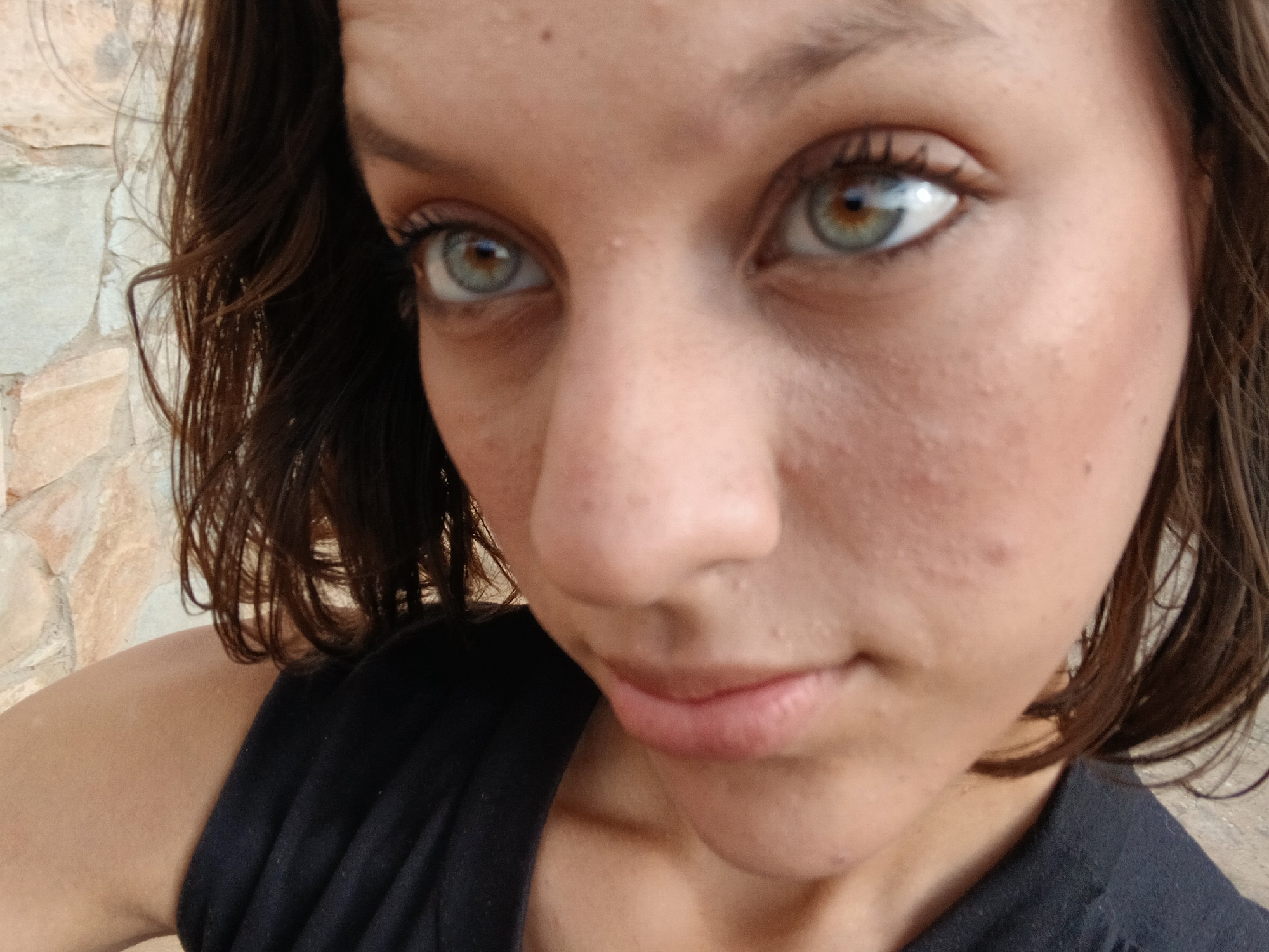 This is a close-up, eye-level, medium shot of a young woman's face, cropped from the chest up, with the camera positioned slightly above her eye level, looking down at her. She has fair skin with visible pores and a few scattered blemishes. Her eyes are large and a striking greenish-brown color with dark eyelashes. Her eyebrows are dark and naturally shaped. Her nose is prominent and slightly upturned, and her lips are full and pale pink. Her dark brown hair is slightly wavy and falls around her face and shoulders. She is wearing a black, sleeveless top. The background is a rough, textured stone wall in a muted earthy tone. The image is well-lit, suggesting an outdoor setting, likely in Santa Ana, Venezuela, where the warm climate and architecture are consistent with this backdrop. The focus is sharp on her face, with a slight blur in the background, creating a shallow depth of field.