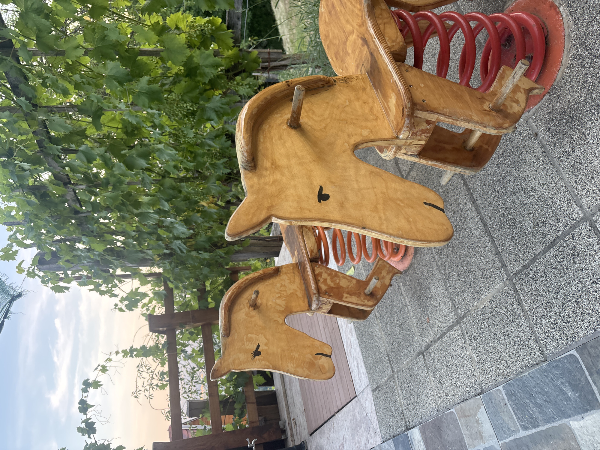 The image depicts two wooden rocking horses with red springs, set on a tiled patio. The horses have simple painted eyes and mouths. In the background, a wooden fence and lush green grapevines are visible. The sky above is a pale blue with wispy clouds, suggesting either early morning or late afternoon. The scene appears to be an outdoor recreational area, possibly a garden or backyard, with elements of nature and play. The specific location is identified as Dolenje Grčevje, Slovenia.