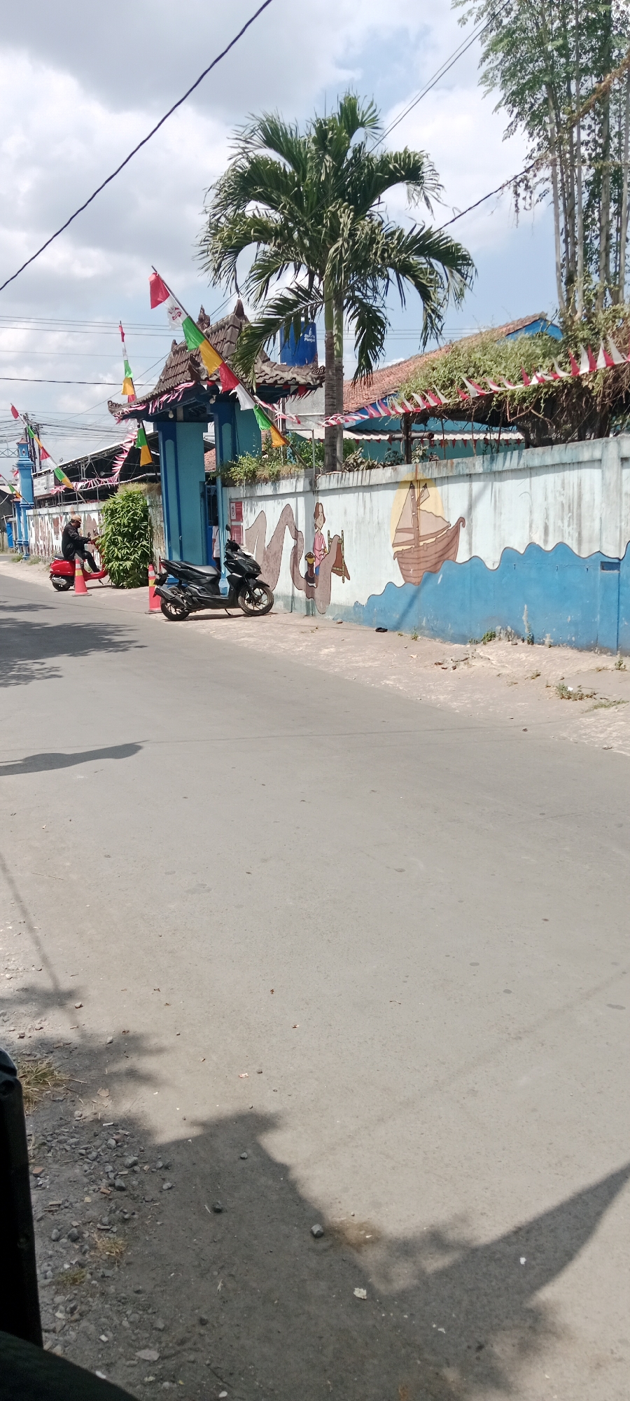 In a sunny outdoor scene, a paved road runs alongside a blue and white painted wall adorned with a mural. The mural depicts a person riding on a mythical creature, possibly a mermaid or serpent, with a sailboat in the background, set against a yellow sun and wavy blue water. To the left of the wall is an entrance gate with a traditional roof, decorated with colorful triangular flags. Two motorbikes are parked on the side of the road in front of the wall and gate. One is a black scooter, and the other is a red scooter with a person wearing a black jacket and helmet riding it. The road itself is gray asphalt with some scattered gravel and debris. Palm trees and bamboo trees are visible in the background, suggesting a tropical or subtropical environment. The sky is partly cloudy, with white and gray clouds. The overall impression is a street scene in an Indonesian city, likely Purwomartani, Indonesia, given the architectural style and decorations.