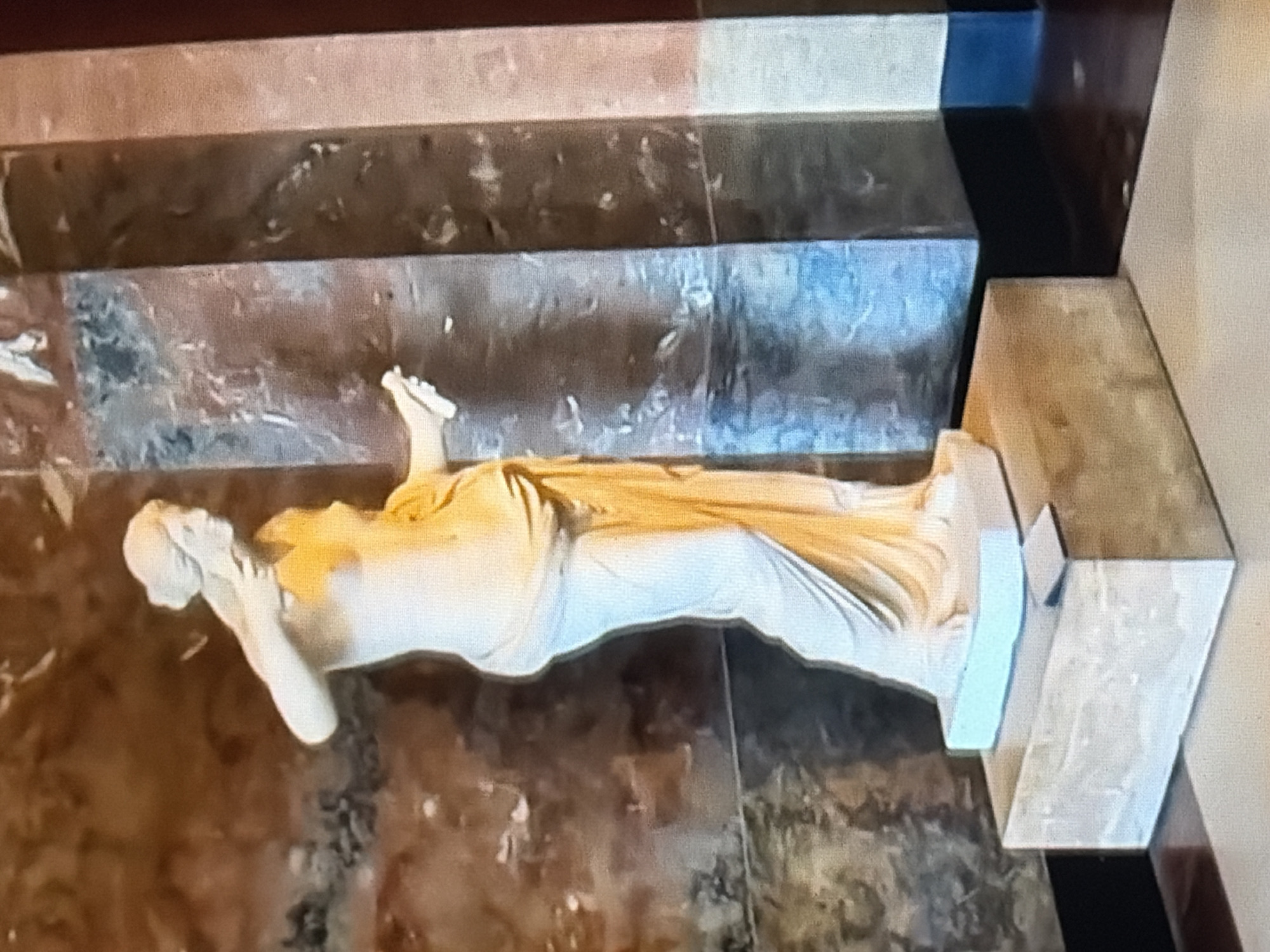 A life-sized, pale sculpture of a woman is positioned on a light brown, rectangular pedestal in the center of the frame. The sculpture depicts the woman in a flowing, toga-like garment, with her left arm raised and her right hand extended, holding a scroll. The background features a large, textured pillar with marbled patterns in shades of brown, blue, and white, and a wall with similar brown marbling. The floor is a polished surface reflecting the light. The scene appears to be indoors, likely in a museum or gallery.