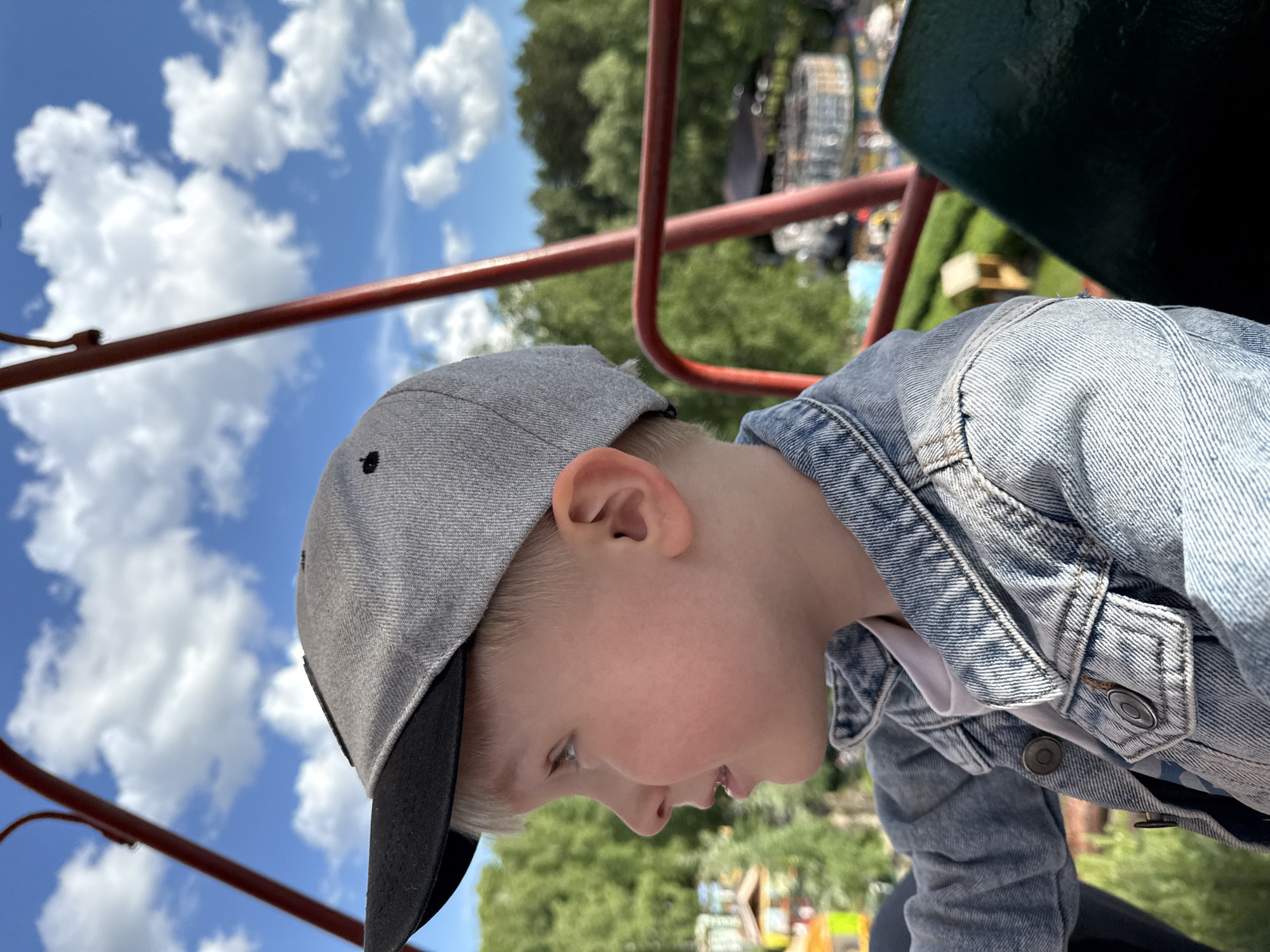A young child wearing a gray baseball cap and a denim jacket is seen from a low angle, seemingly on a ride, possibly a Ferris wheel, in a park. The backdrop includes green trees and a blue sky with scattered white clouds. Red metal bars and the edge of a dark green seat are visible, suggesting a fairground or amusement park setting. It's impossible to confirm this is specifically in the city of Chaikovsky, Russia, based only on the information of this image.