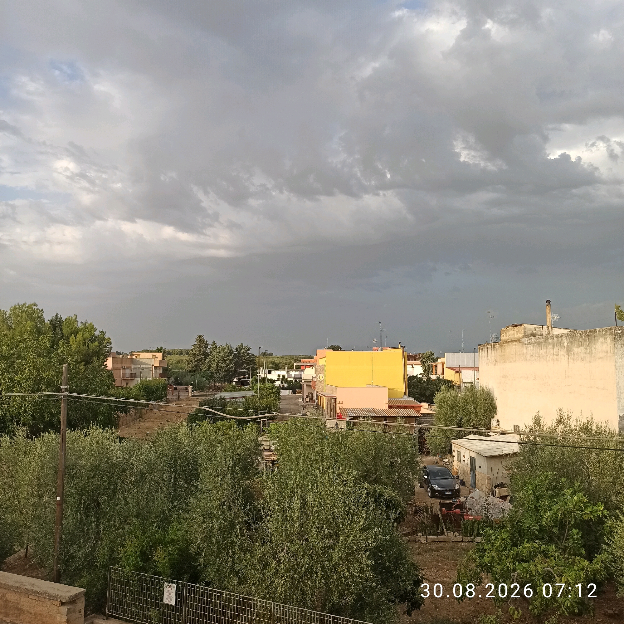 The image is an outdoor, slightly elevated view of a residential area with a cloudy sky. In the foreground, there is a fence and some vegetation. In the middle ground, there are several buildings, some painted in bright colors like yellow and pink, and others with a more muted beige tone. Trees and smaller structures are interspersed among the buildings. A black car is parked in front of a small building. The background shows more buildings and vegetation, with a hilly landscape in the distance. The date and time stamp "30.08.2026 07:12" is visible in the lower right corner. The location is specified as Bitonto, Italy.