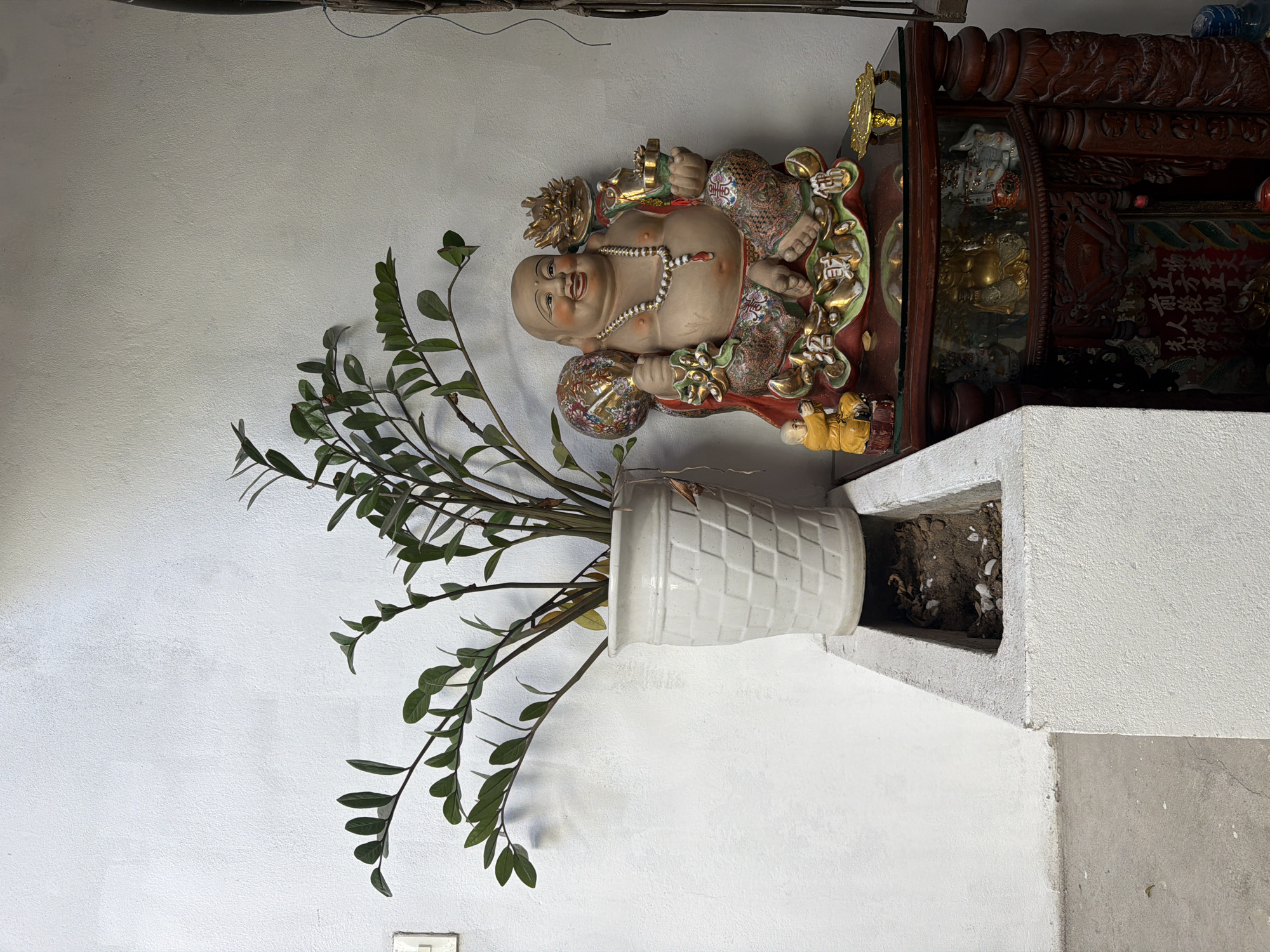 A ceramic statue of a smiling, rotund figure, adorned with a beaded necklace, sits on a dark wooden table in front of a white textured wall. The statue is richly decorated with colorful patterns on its clothing and holds various symbolic objects in its hands, including a round, patterned sphere and a small golden vessel. To the left of the statue, a tall white pot holds a green leafy plant. In front of the pot is a concrete planter box containing soil and some debris. The table also displays a small yellow figurine of a person kneeling. The overall scene suggests an indoor setting, possibly a home or a shop, given the decorative elements and the placement of the items. The image does not provide specific visual cues to confirm its location within Hoi An, Vietnam.