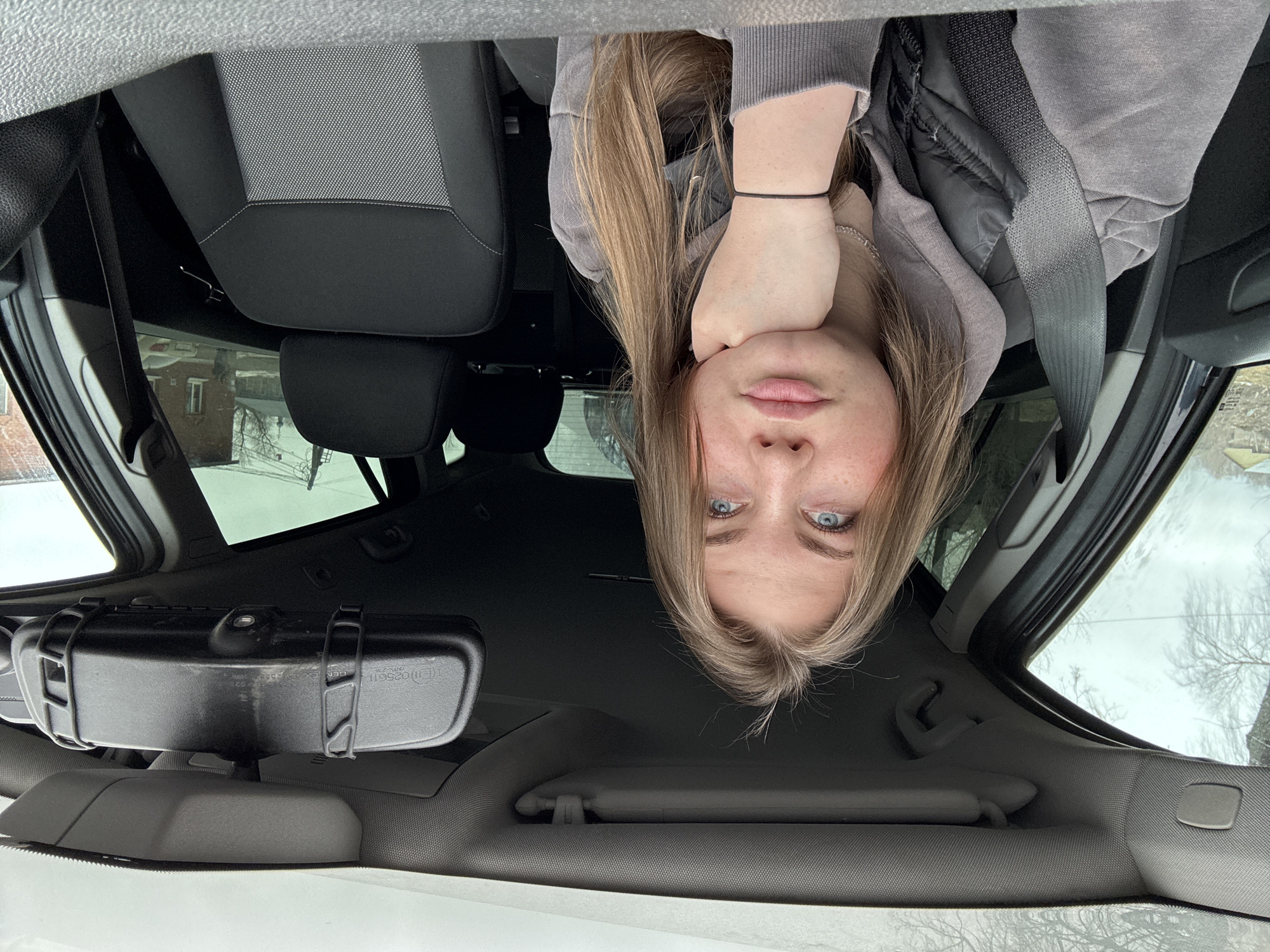 A woman is sitting in the driver's seat of a car, facing the camera, which is oriented upside down. She has fair skin, light brown hair, and blue eyes. She is wearing a gray hoodie, a black vest, and a seatbelt. Her left hand is resting on her face.
The car interior is visible, including the gray and black seat, the rearview mirror with a black case attached to it, the sun visor, and the car's roof. Outside the car windows, bare trees and building are visible. Based on this, the location is in the city of Фоки, Russia.