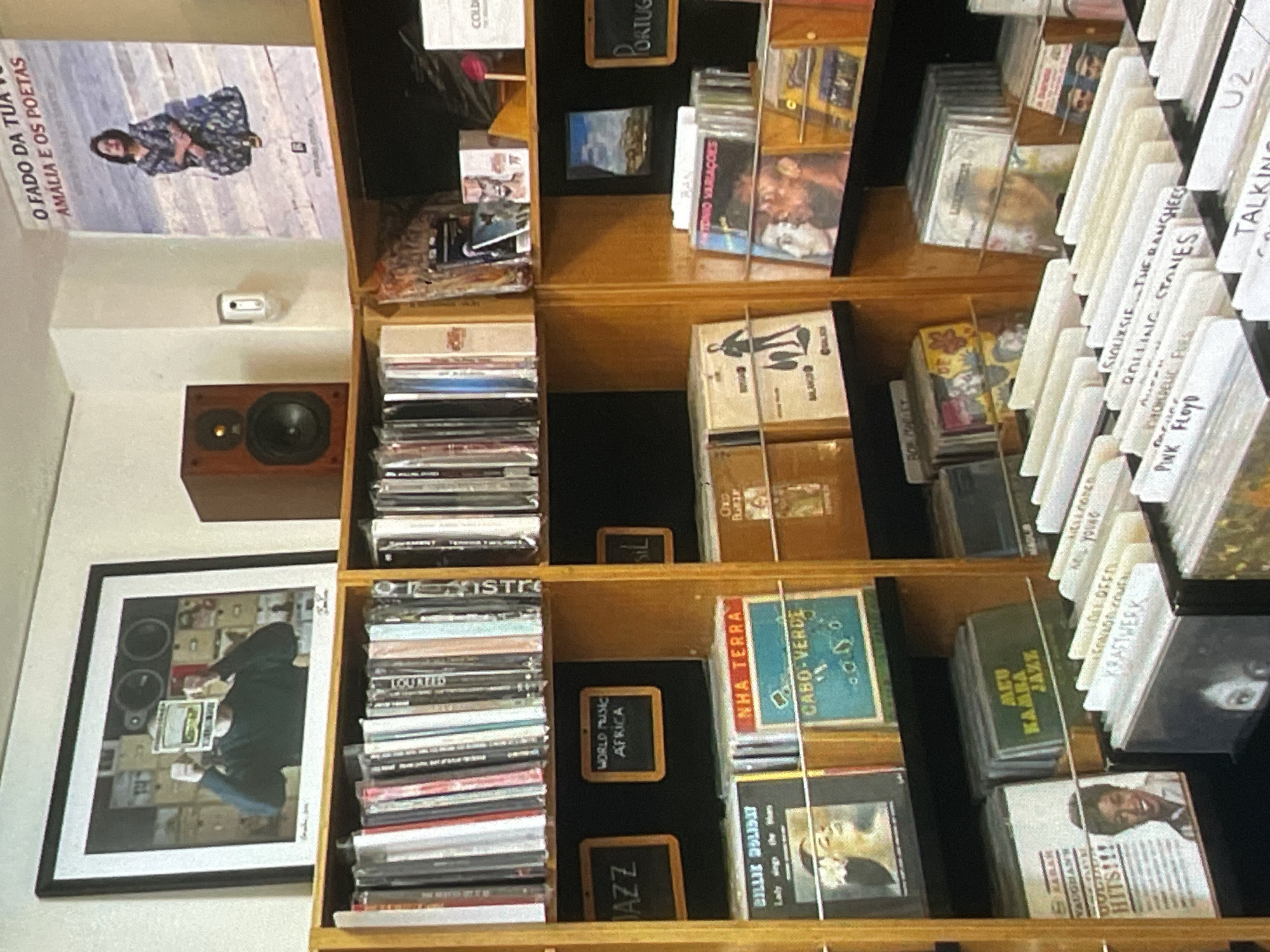 The image depicts the interior of a record store. The store features wooden shelves filled with vinyl records and CDs. There is a framed picture on the wall above the shelves, and a speaker is positioned on a shelf. A poster is also visible on the wall. Several signs are placed on the shelves, indicating genres or origins of the music, such as "JAZZ", "WORLD MUSIC AFRICA", and "PORTUGAL". The records and CDs are neatly organized, with some cases displaying album art and artist names. In the foreground, there are rows of records with visible labels. The lighting appears to be artificial. There are no visible people in the image, and no specific details about the city of Harmanli, Bulgaria, are discernible from the visual content.