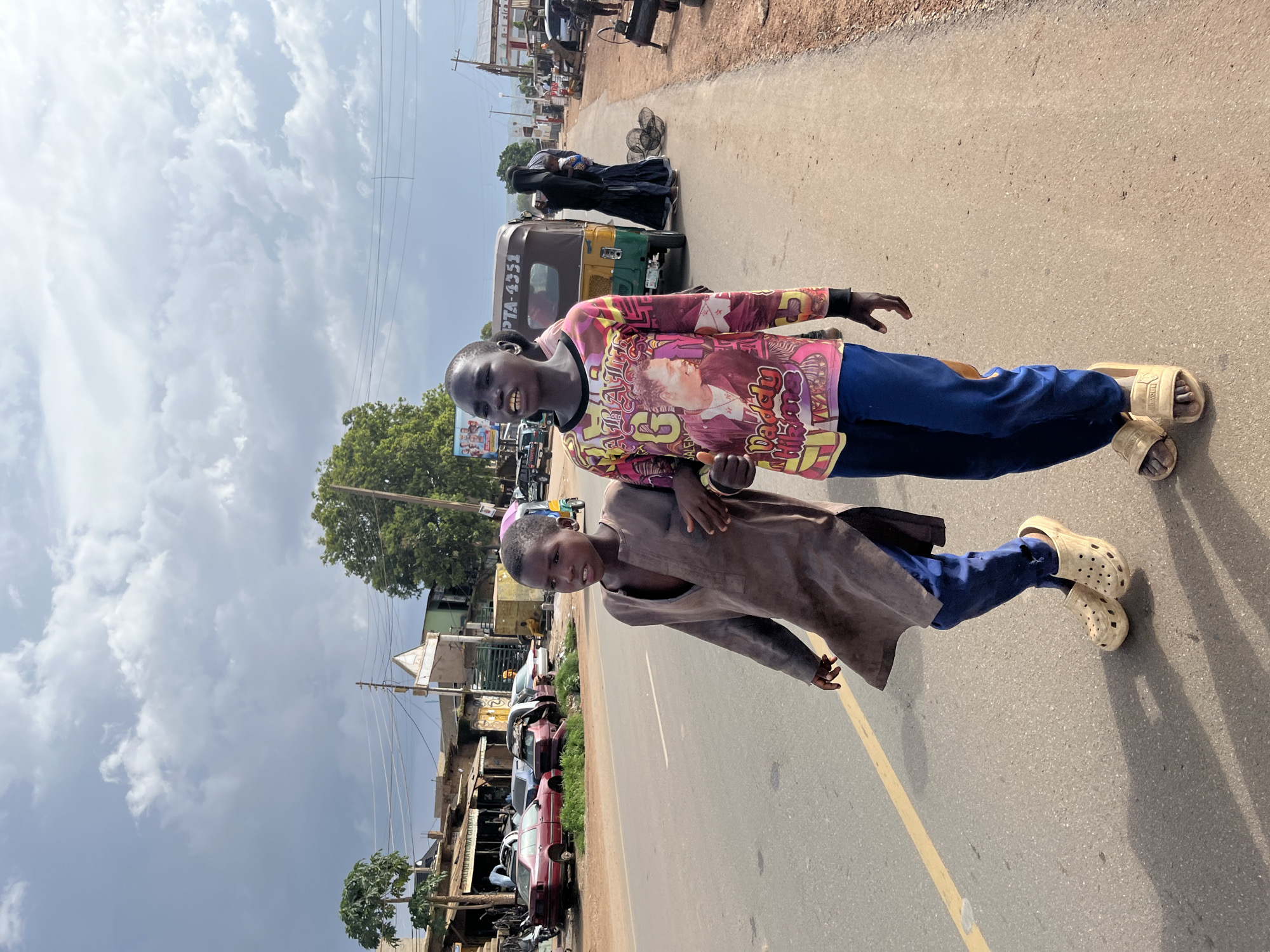 Two young boys walk on the side of a paved road in Jos, Nigeria, under a cloudy sky. The boy on the left wears a loose, long-sleeved brown tunic and blue pants, with tan clogs on his feet. The boy on the right wears a colorful long-sleeved shirt with graphics and blue pants, also wearing tan clogs. They walk side-by-side, appearing to be interacting. In the background, several cars are parked along the road, some appearing older or damaged. A yellow and green auto-rickshaw is visible, along with a person wearing a black hijab and long dress. Buildings line the roadside, some with corrugated metal roofs and awnings. The ground on the right side of the road is unpaved and appears to be reddish-brown dirt. Power lines and utility poles are present. The overall scene depicts a bustling street environment with a mix of vehicles, buildings, and pedestrian activity.