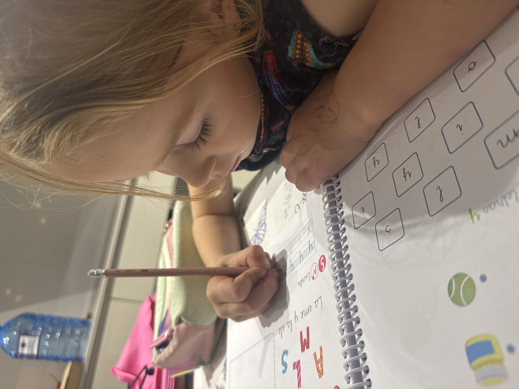 A young child with fair hair is diligently working on a workbook. The child is bent over the open book, holding a pencil in their right hand, carefully writing in it. The book appears to be an educational exercise book, featuring handwriting practice. The background includes elements of a home environment, such as a water bottle and a pink backpack or bag. The image does not contain enough information to determine if it is in Valencia, Spain.