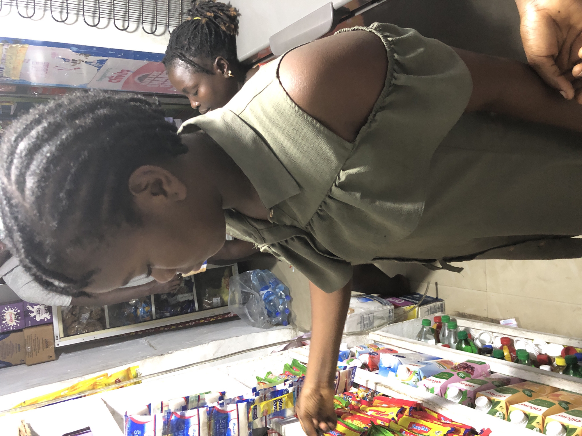 A girl is reaching for snacks on a shelf in a store. The girl has her hair in braids and is wearing a green shirt with her shoulders exposed. Behind her, a younger child is leaning against a counter. The shelves are stocked with various food items, including packaged snacks, bottles of water, and cartons of juice. The store appears to be a small, local shop. The image is a close-up of the girl and the immediate surroundings, with the background blurred. The scene is lit by artificial light, suggesting it is indoors. The overall impression is one of a casual shopping trip in a bustling neighborhood market.