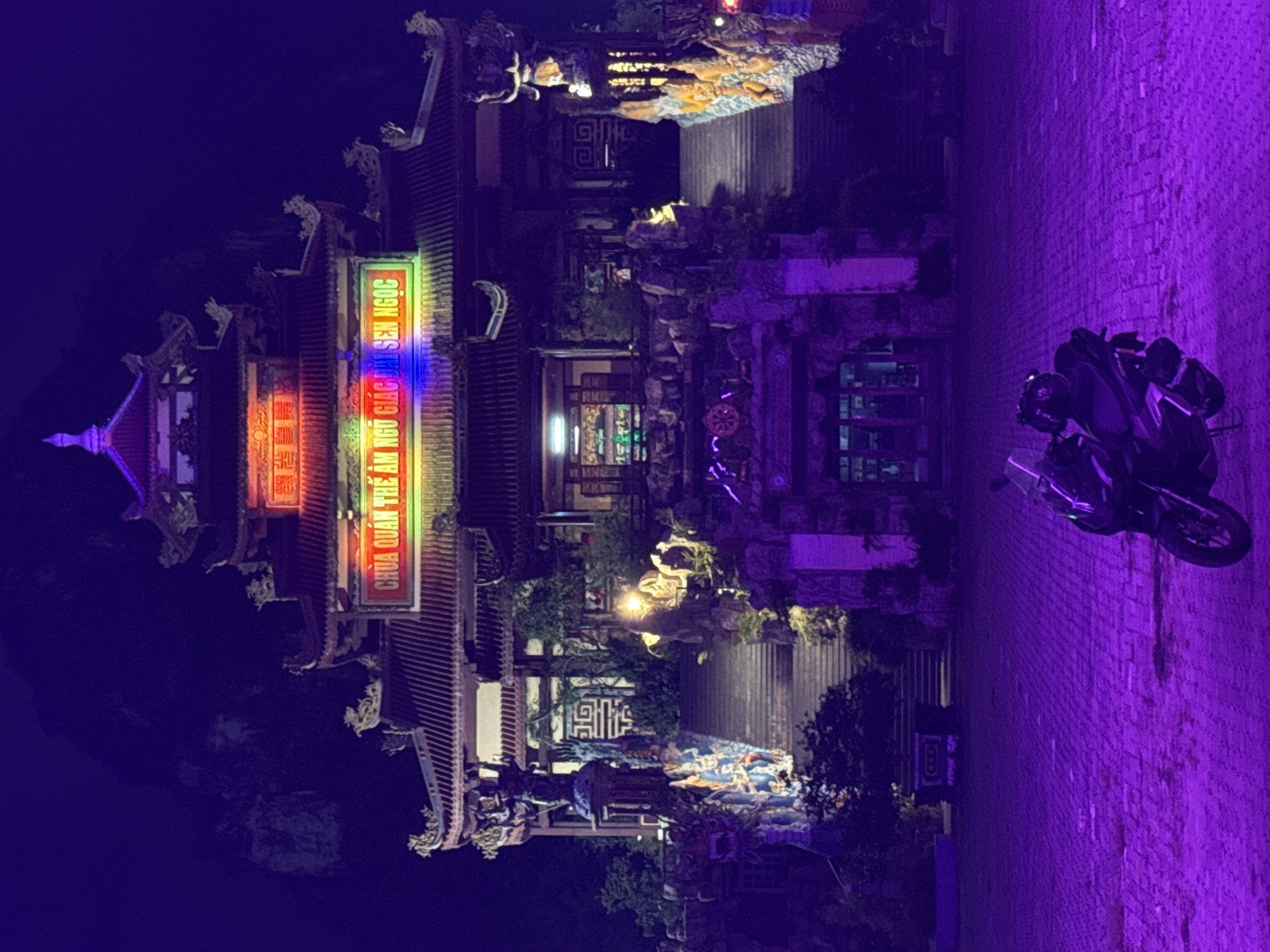 Here is a description of the image:
The image depicts the illuminated Buddhist temple of Chùa Quán Thế Âm, in Đà Nẵng, Vietnam, at night. The temple's architecture is traditional Vietnamese, featuring tiered roofs with ornate decorations. Colorful neon signs with Vietnamese text are visible on the temple's facade. There are detailed rock gardens and landscaped areas around the temple. A parked motorcycle is visible in the right of the image. The entire scene has a purple or blue hue due to the lighting conditions.
