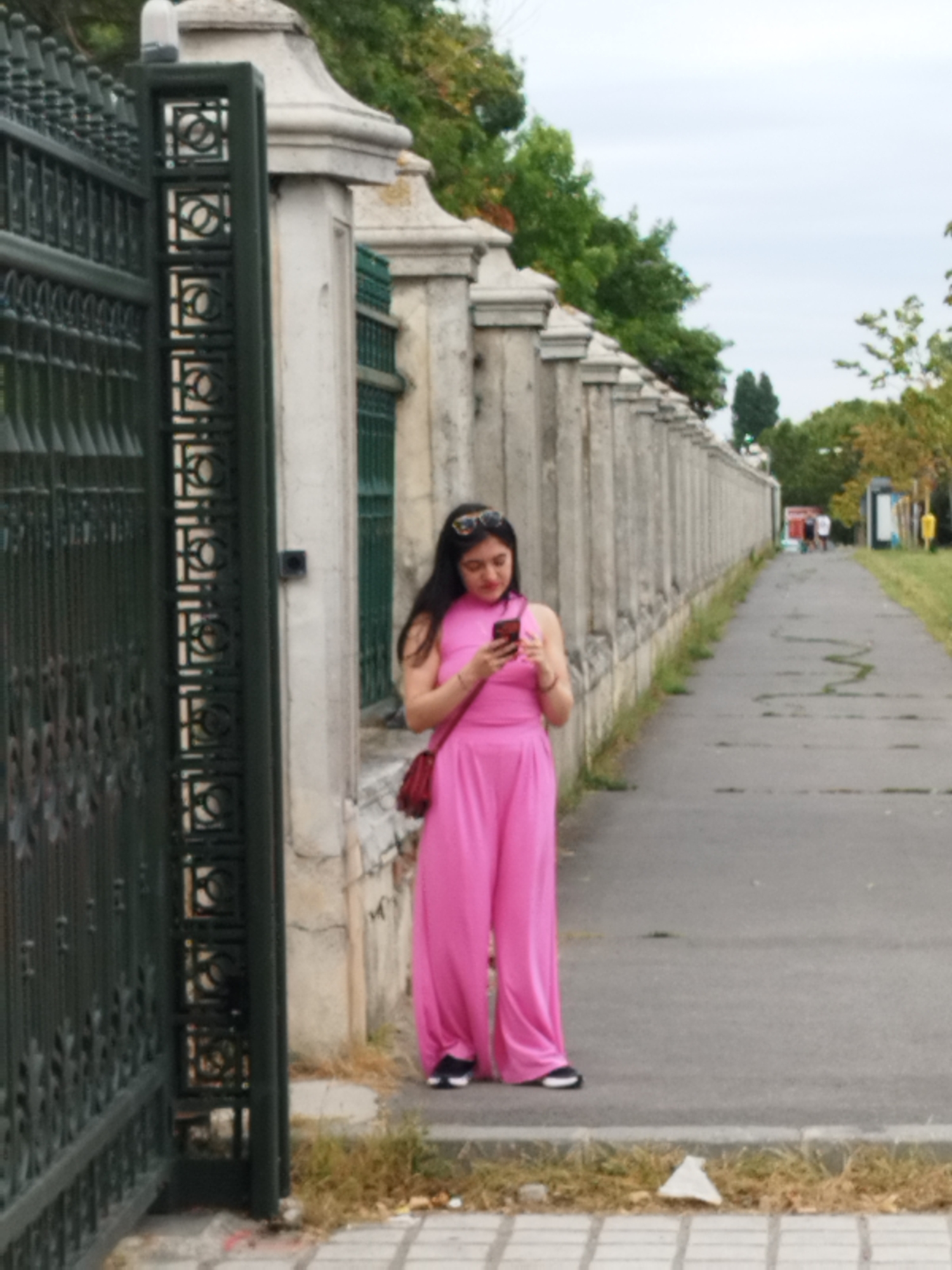 A young woman in a bright pink, sleeveless top and wide-leg pants stands on a paved pathway next to a stone wall. She is looking down at her phone, which she holds in her right hand. She has long, dark hair and is wearing sunglasses on her head. A dark red crossbody bag is slung over her left shoulder. To her left is a tall, dark green metal fence with intricate geometric patterns. The pathway continues into the distance, flanked by the stone wall on one side and a grassy area with trees on the other. In the background, other people are visible walking along the pathway, and there are various streetlights and signs. The sky is overcast. The scene appears to be a public park or walkway within a city. This location is the park of the Palace of the Parliament in Bucharest, Romania.