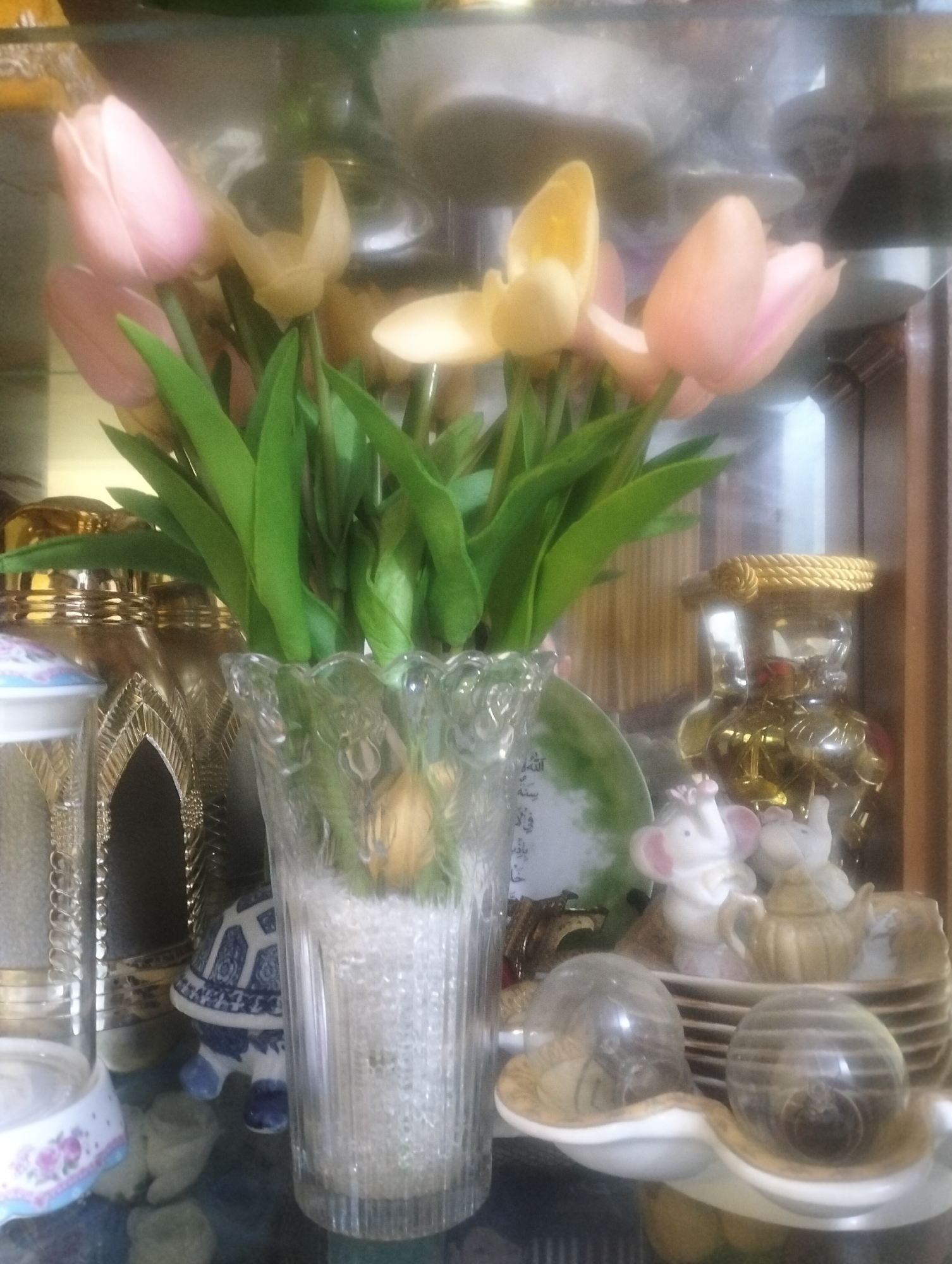 The image depicts a shelf display with a floral arrangement and various decorative items. In the foreground, a clear glass vase contains a bouquet of pink and yellow tulips with green stems and leaves. The vase is filled with small, clear, bead-like objects. To the left of the vase, a tall, ornate golden container with intricate patterns is visible, partially obscured by a white and floral patterned object. Behind the vase, a decorative plate with a green hue and Asian calligraphy is displayed.
To the right of the vase, a stack of decorative plates is topped with two small, white elephant figurines, one with pink ears and a pink flower on its head, and a small, tan teapot. Further to the right, another ornate golden container with a twisted rope-like design is prominent. In front of the plates and elephant figurines, two clear glass domes, each containing a decorative item that appears to be a ball or orb, rest on white ceramic dishes. Below the elephant figurines and teapot, a blue and white patterned ceramic turtle figurine is positioned.
The overall setting suggests an indoor display, possibly in a home or shop, characterized by a collection of decorative objects. The lighting is soft and diffused, creating a slightly hazy effect. The background appears to be a reflective surface, possibly glass or polished wood, with further items visible in the distance, but they are out of focus. The location within Dumai City, Indonesia, is not explicitly indicated by any visual cues in the image itself.