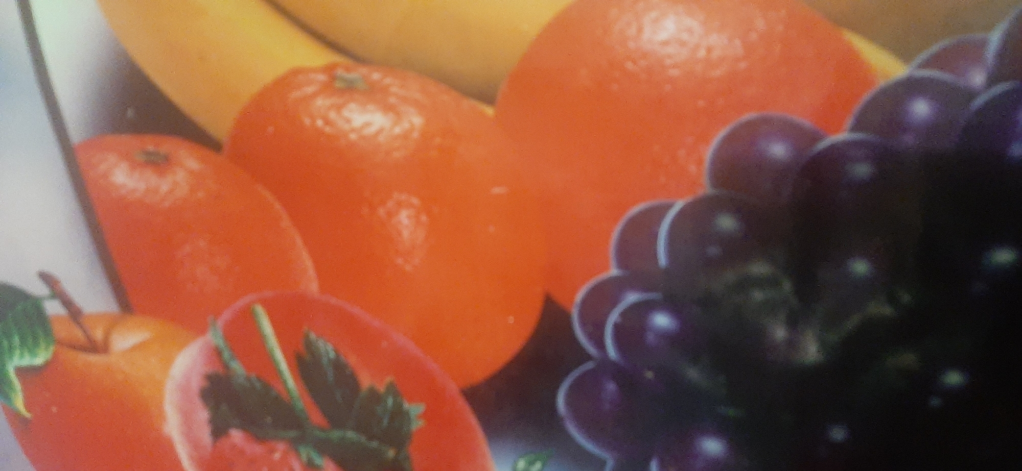 The image is a close-up, high-angle, indoor photograph of a fruit still life. The subjects are fruits, including what appear to be tangerines or clementines, an apple, and a cluster of dark grapes. The fruits are arranged in a bowl or on a surface, though the container is not fully visible. The lighting is soft and diffused, creating gentle highlights on the fruits' surfaces. The background is out of focus and appears to be a light-colored surface, possibly a tablecloth or countertop. There is no indication of the location in Yessentukskaya, Russia, as the image lacks any identifiable landmarks or context related to that specific place.
