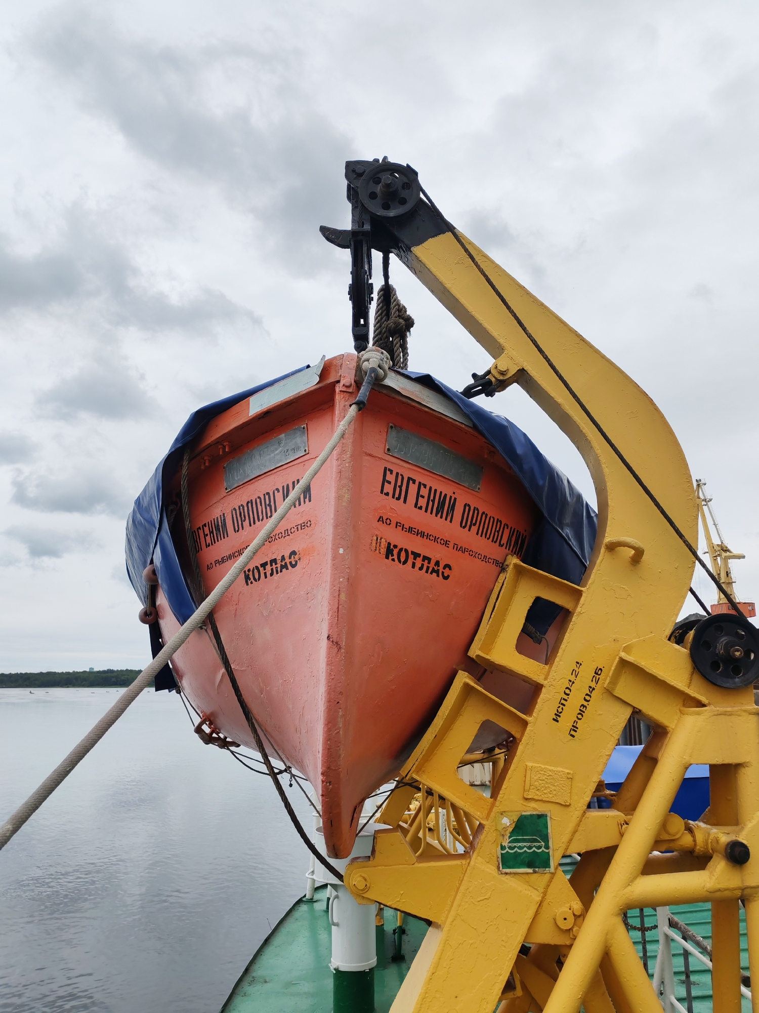 In Bol'shie Mkhi, Russia, a bright orange lifeboat is suspended from a yellow davit crane against a cloudy sky. The lifeboat, partially covered by a blue tarp, bears Cyrillic lettering that reads "Евгений Орловский" and "КОТЛАС". A thick rope is attached to the lifeboat and secured to the deck of a vessel, which is visible in the foreground with its green railing. The crane, a prominent yellow structure, is connected to the lifeboat by cables and pulleys. The scene suggests a maritime setting, possibly a port or a ship at anchor, with calm water and a distant shoreline visible.