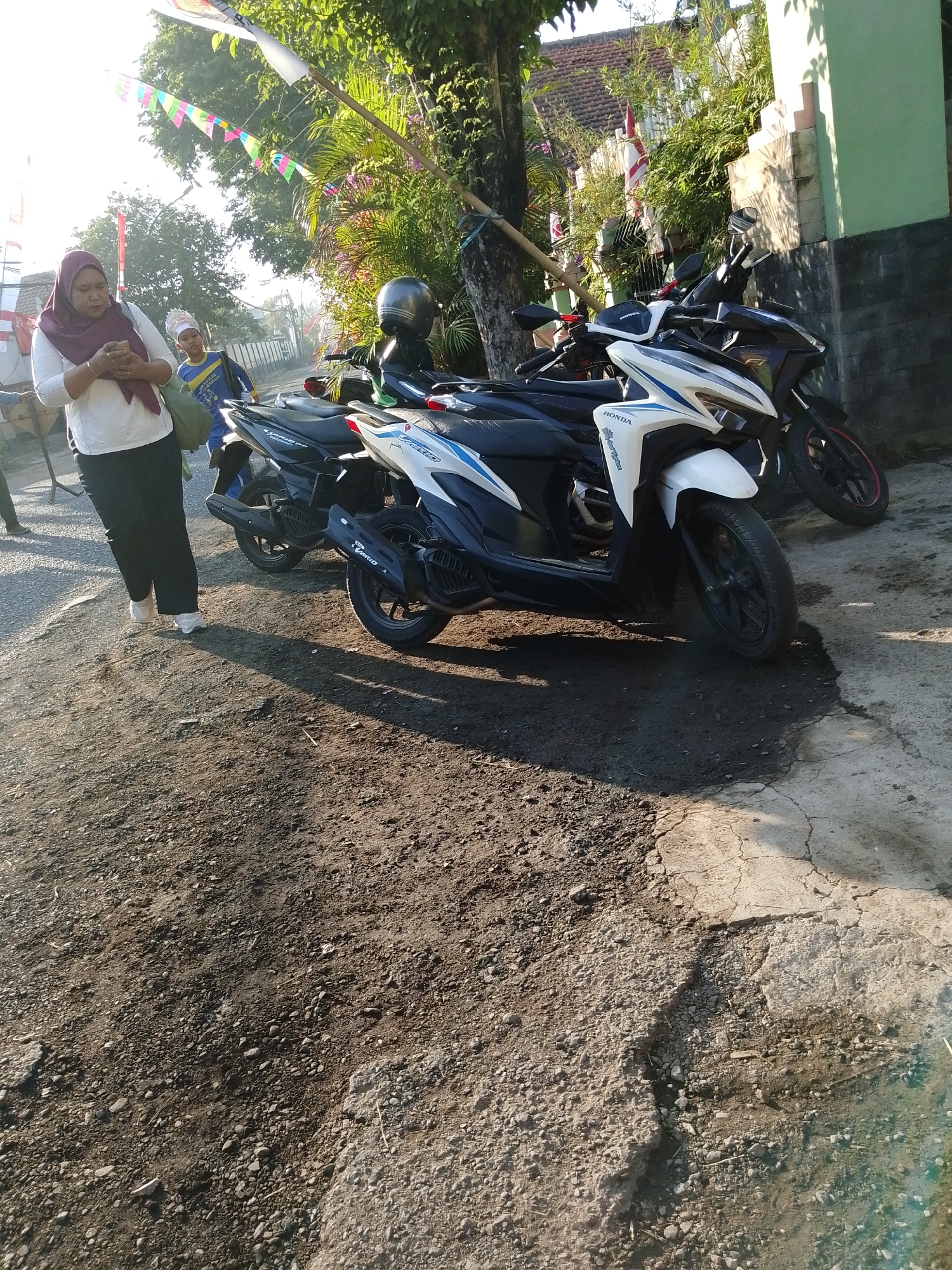 A woman wearing a maroon hijab and a white long-sleeved shirt, paired with black pants and white sneakers, stands on a dirt and gravel path. She is looking down at her phone. Beside her, a young boy in a blue shirt and white head covering walks away from the camera. Behind them, parked along the side of a road, are several motorcycles. Two are prominent in the foreground: one is white with blue accents and the other is dark colored with red accents on the wheel. In the background, there are trees, a building with green and white walls, and colorful triangular flags strung across the scene. The sun is casting long shadows, indicating it is either early morning or late afternoon. This scene appears to be a street in the city of Pasuruan, Indonesia, possibly near a residential area or a school, given the flags and the presence of children.