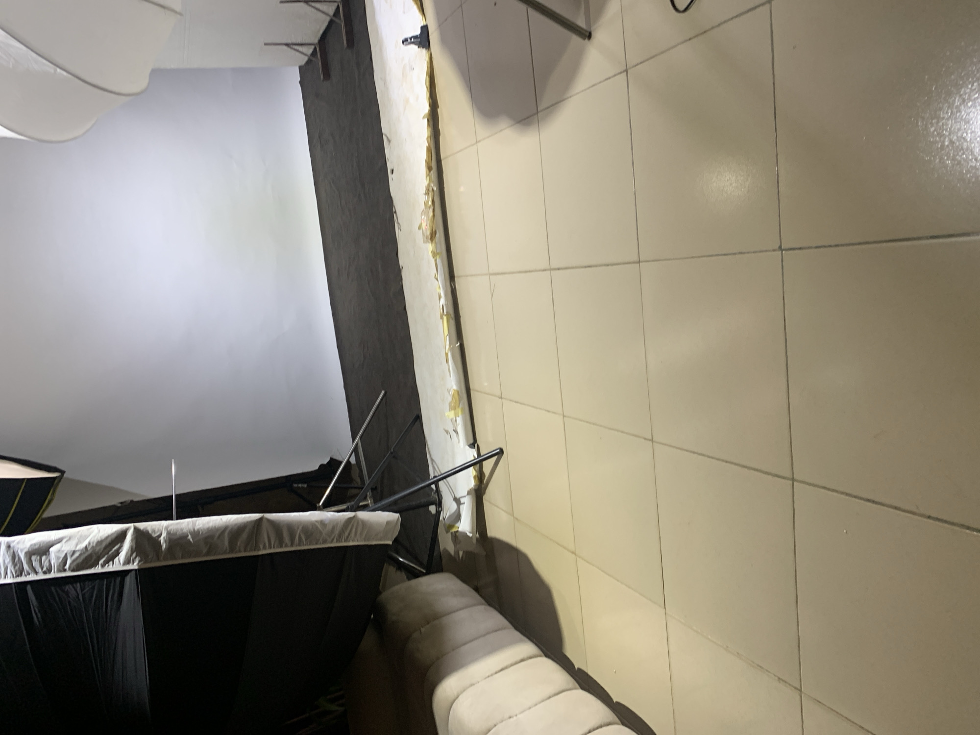 The image depicts an indoor scene that appears to be a studio or a room with a backdrop. The primary subjects are studio equipment, including a large white softbox diffuser at the top left, and what seems to be a backdrop or screen. A dark, cylindrical object with a white rim, likely another piece of studio equipment, is visible in the foreground. Metal stands and frames are also present, suggesting a setup for photography or videography.
The setting is a room with tiled walls and floor. The tiles are a light, neutral color, and the grout lines are visible. A portion of a wall shows peeling paint or wallpaper, revealing older layers underneath, indicating a less-than-perfectly maintained area. A beige-colored sofa or bench with tufted cushioning is positioned near the tiled wall, casting a shadow on the floor.
The lighting suggests that artificial light sources are in use, typical of a studio environment. The overall mood is functional, geared towards creative work. There is no indication of people or specific activity beyond the setup of the equipment itself. The time of day and weather are not discernible from the image. No visible text is present in the frame.
The location context of Barakin Sabawa, Nigeria, is provided, but the visual elements within the image do not offer specific cues to confirm or deny this location. The scene is characterized by its utilitarian nature, focusing on the tools and materials for production.