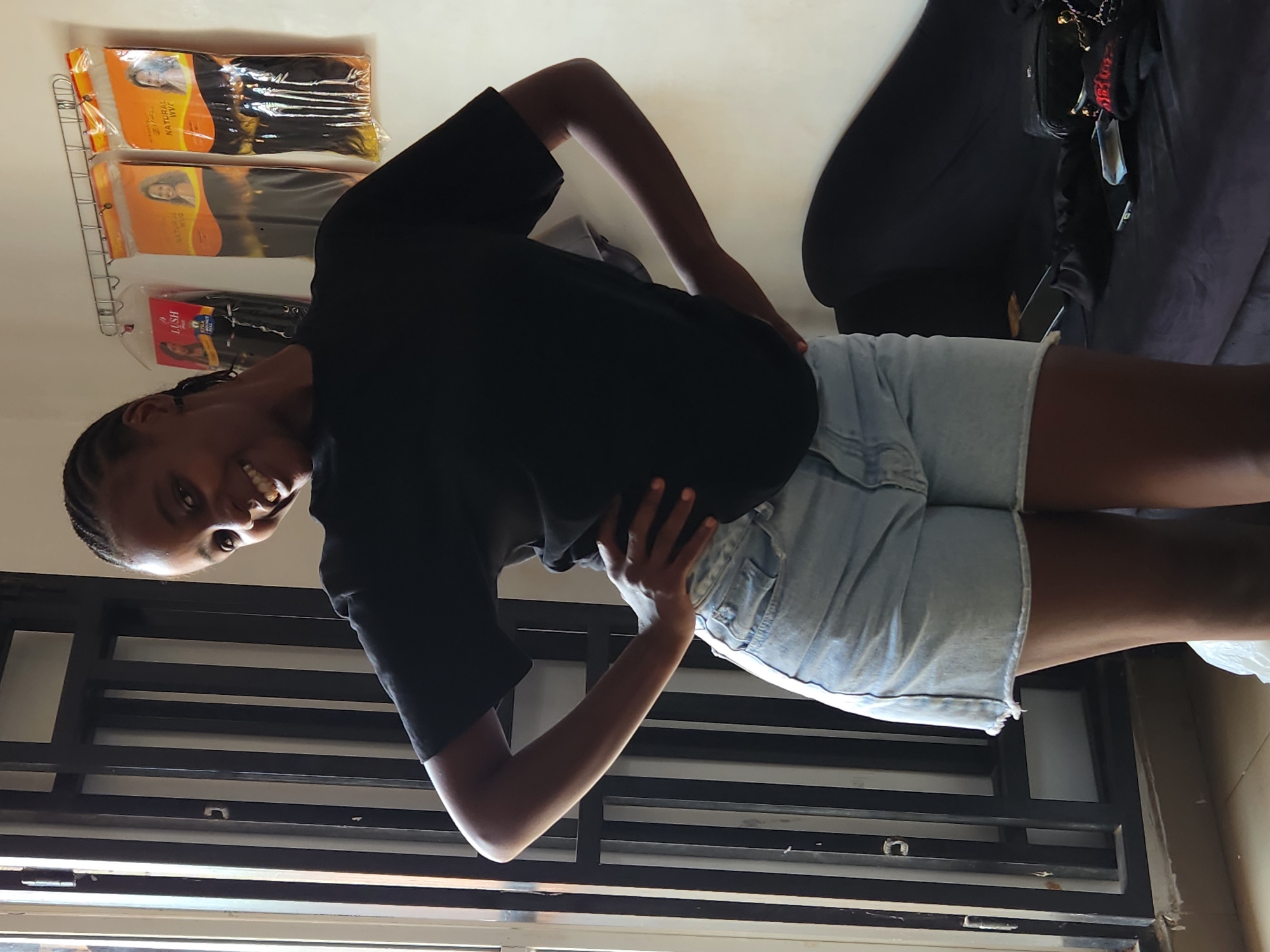 A Black woman with braided hair stands with her hands on her hips, smiling at the camera in Sabon Tasha, Nigeria. She is wearing a black t-shirt and light-wash denim shorts. Behind her, on the right, a dark velvet chaise lounge is visible with several handbags and cell phones scattered on it. To the left of the woman, a black metal gate is present. On the wall behind her, several packages of hair extensions are hanging from a wire rack. The lighting suggests it is daytime, with natural light coming from the left side of the image.