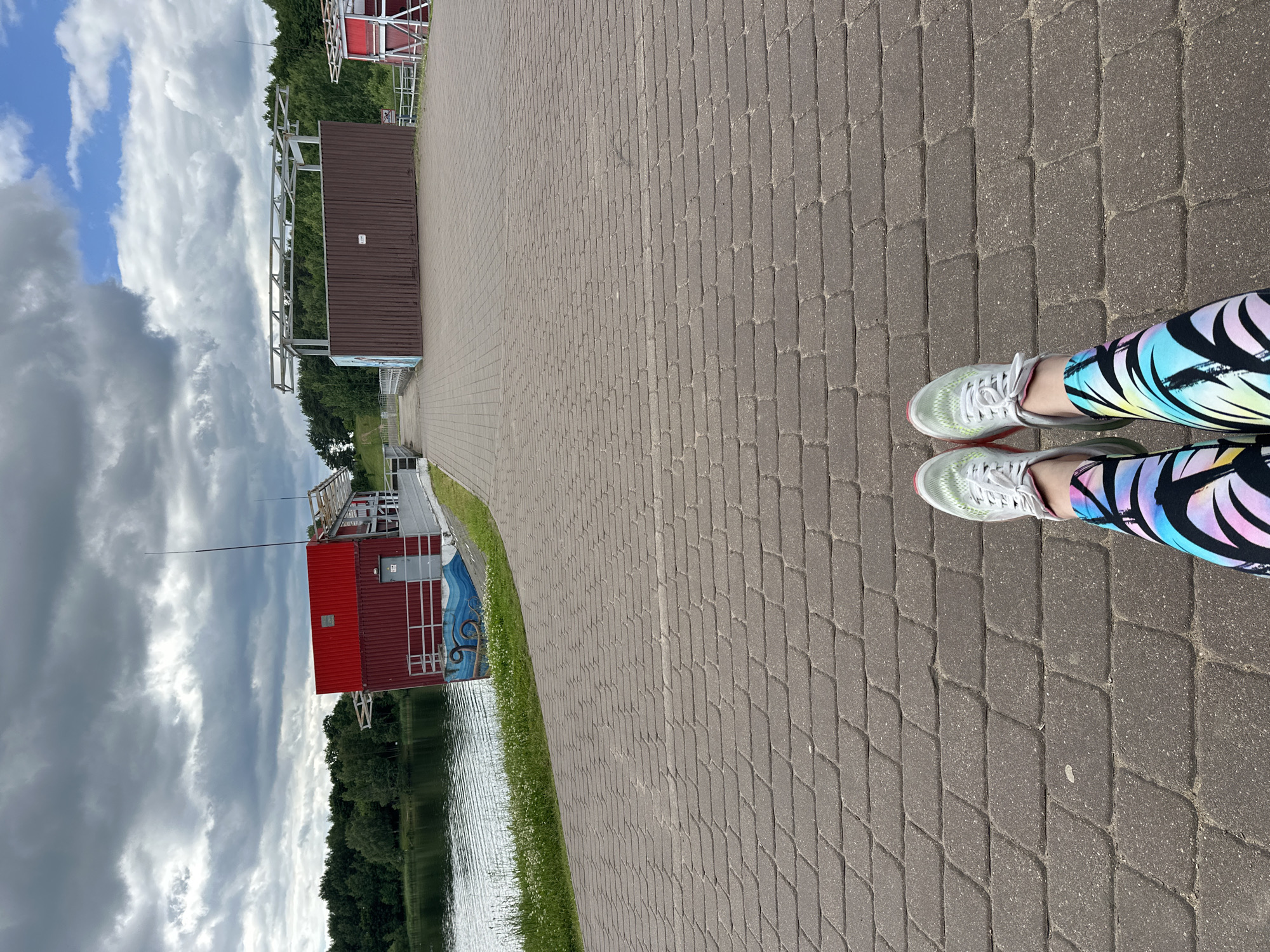 The image depicts a person's legs from the knees down, standing on a paved walkway next to a body of water. The person is wearing athletic shoes and colorful leggings with a pattern of abstract brushstrokes in shades of blue, yellow, pink, and black. The walkway is made of interlocking pavers, and a thin strip of green grass separates it from the water.
In the background, a red building with a metal frame structure on top is situated on the edge of the water. This building appears to be a boathouse or a similar facility. Beyond the building, a dense line of green trees forms the backdrop, suggesting a natural or park-like setting. The sky is filled with dramatic, dark clouds, indicating a possibly overcast or stormy day. A thin pole, possibly a flagpole or a part of an antenna, extends vertically from behind the red building into the sky.
The image is oriented sideways, as indicated by the vertical orientation of the subjects and the environment. The scene suggests an outdoor activity, likely walking or running, given the attire of the person. The weather appears to be unsettled, with a mix of dark clouds and glimpses of lighter sky. The location cues, such as the paved walkway, the water, the boathouse, and the trees, are consistent with a park, waterfront recreation area, or a sports facility. No visible text is present in the image.