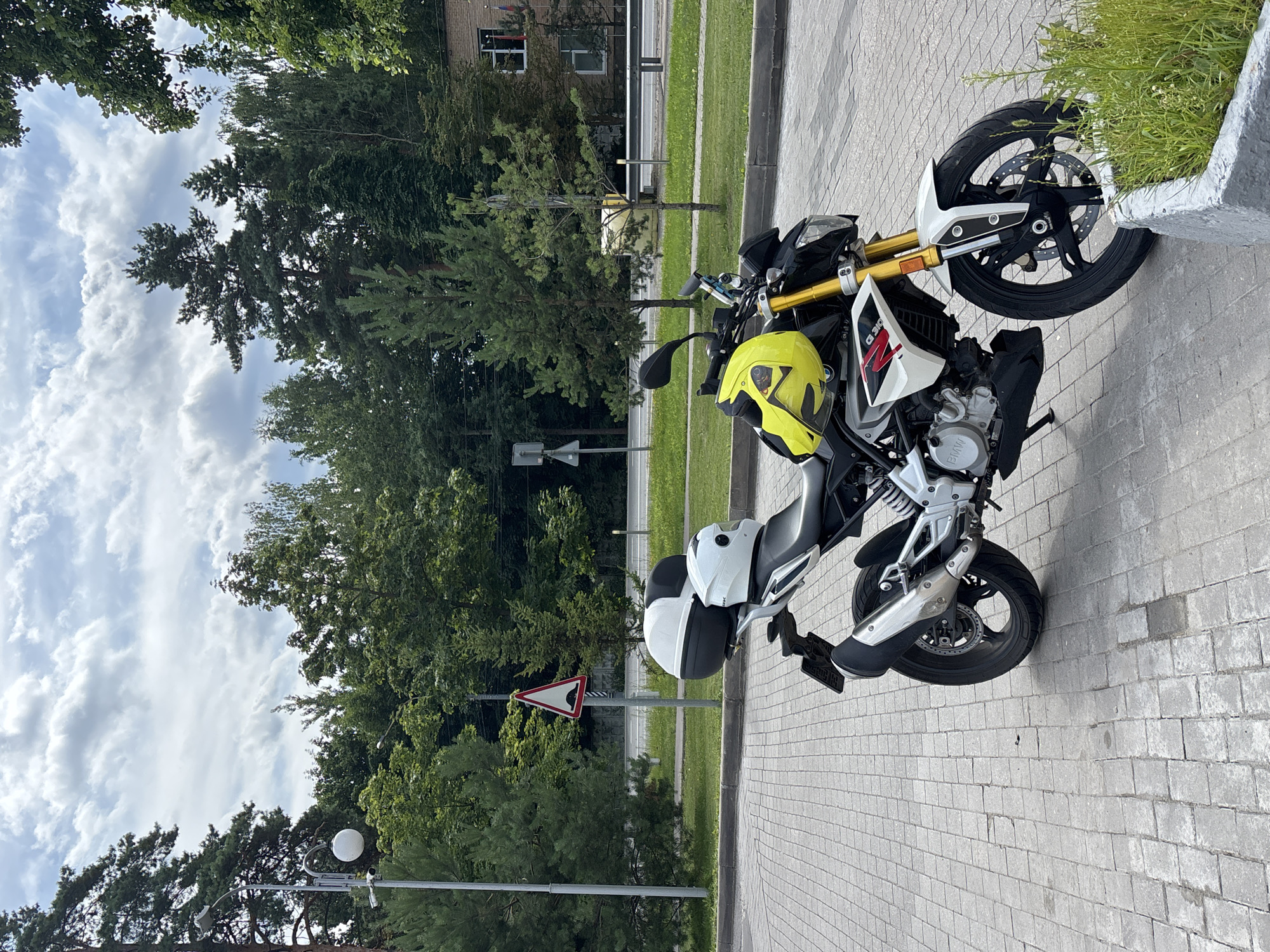 A white and yellow BMW G 310 R motorcycle with a black top case and a white helmet rests on a paved surface in front of a line of trees and a road. A speed bump sign is visible behind the motorcycle. The sky is overcast with scattered clouds. The scene appears to be in a park or roadside area in Gorki-2, Russia.