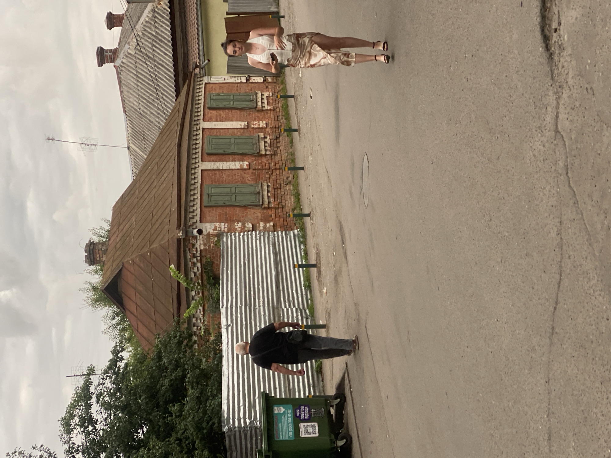 An elderly Caucasian male with white hair wearing a black t-shirt, dark pants, and a black crossbody bag walks away from the camera on a paved street. To his left is a large green dumpster with a QR code and text on it. Behind him is a corrugated metal fence. In the background, there is a brick building with green shutters and a reddish-brown metal roof. To the right of the brick building, a younger Caucasian female wearing a white halter top and a patterned skirt with a slit walks towards the camera, looking down at her phone. She is wearing heeled sandals. The sky is overcast and the environment appears to be a city street in Poltava, Ukraine.