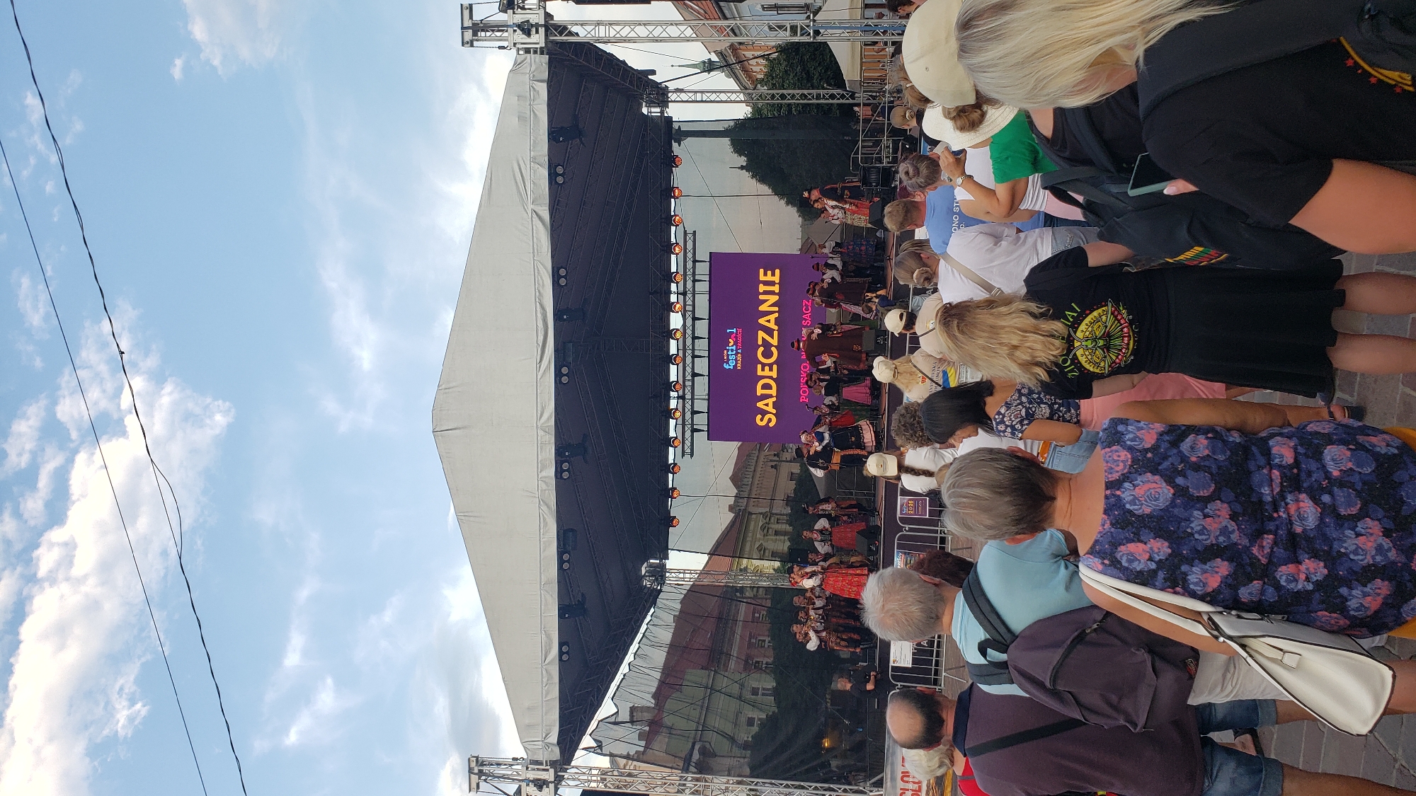 The image depicts an outdoor festival performance on a stage in Prešov, Slovakia. A group of performers in traditional folk costumes are dancing on stage, with a large screen behind them displaying the text "Sadeczanie" and "POLSKO MOJE SACZ". The audience, consisting of dozens of people, stands in front of the stage, watching the performance. The event appears to be taking place during the day, under a cloudy sky.