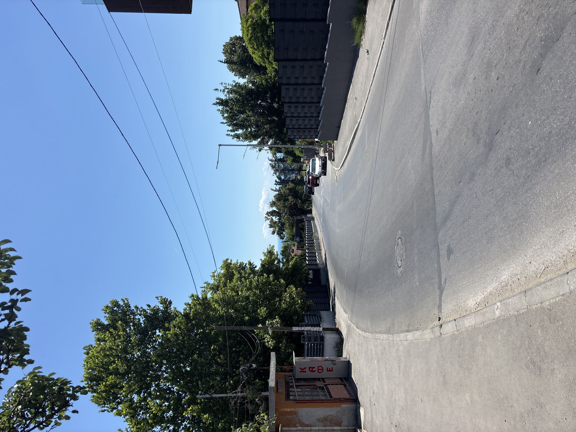 The image depicts a sun-drenched street scene in Varna, Bulgaria, likely during daytime. The road curves to the right, leading uphill. On the left side of the road, there's a small, orange building with a metal gate and the Cyrillic word "КАФЕ" (KAFE) visible, indicating a cafe. A sidewalk runs alongside the building. Mature green trees with dense foliage are prominent on both sides of the street, with some branches extending into the top of the frame. A utility pole with numerous wires stands near the cafe.
Further down the road, several cars are visible: a red SUV, a silver sedan, and another light-colored car. The road is paved and appears to be in moderate condition, with some patches and markings. On the right side of the road, a dark grey, modern fence with a textured pattern lines the street. Beyond the fence, glimpses of buildings and more trees suggest a residential or commercial area.
In the distance, a clear blue sky stretches across the horizon, with faint hints of the sea or coastline visible between the trees. The overall environment suggests a warm, sunny day in a relatively quiet urban or suburban setting within Varna. There are no people visible in the image.