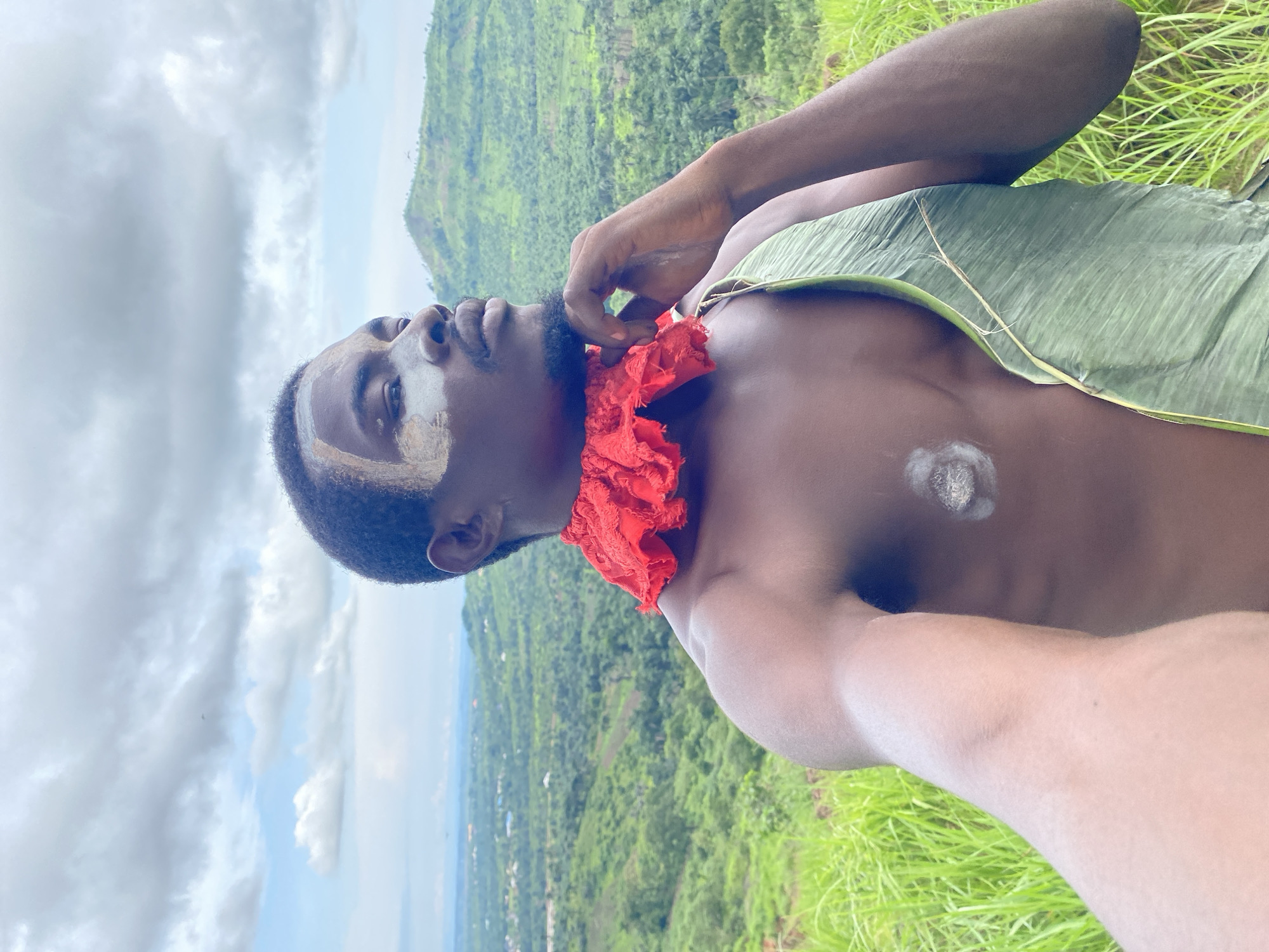 This media file is a portrait of a Black man with a red cloth around his neck and a green leaf draped over his chest. He has white markings on his face and chest. The man is looking upwards with a pensive expression. He is positioned on a grassy hillside with a lush green valley and a village in the background. The sky is cloudy. The scene appears to be set in Ufu Esioni, Nigeria. The overall mood is serene and contemplative, perhaps suggesting a connection to nature or cultural traditions.