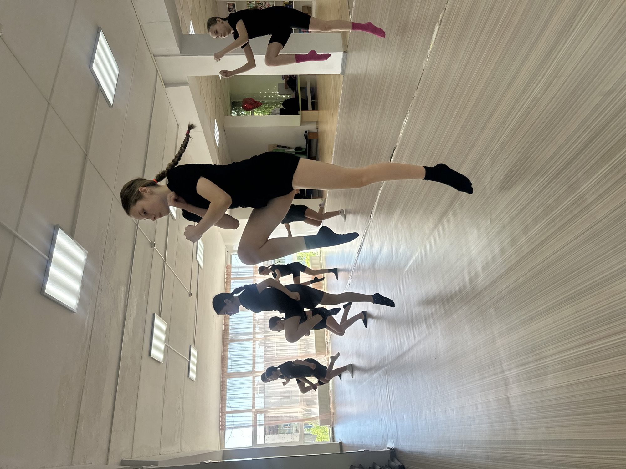 In what appears to be a dance studio or gymnasium, a group of young women are performing a coordinated movement exercise. Most are wearing black shorts and tops with different colored socks. The studio features linoleum floors, white walls, fluorescent lighting fixtures, and windows with semi-transparent curtains. The location is Votkinsk, Russia.
