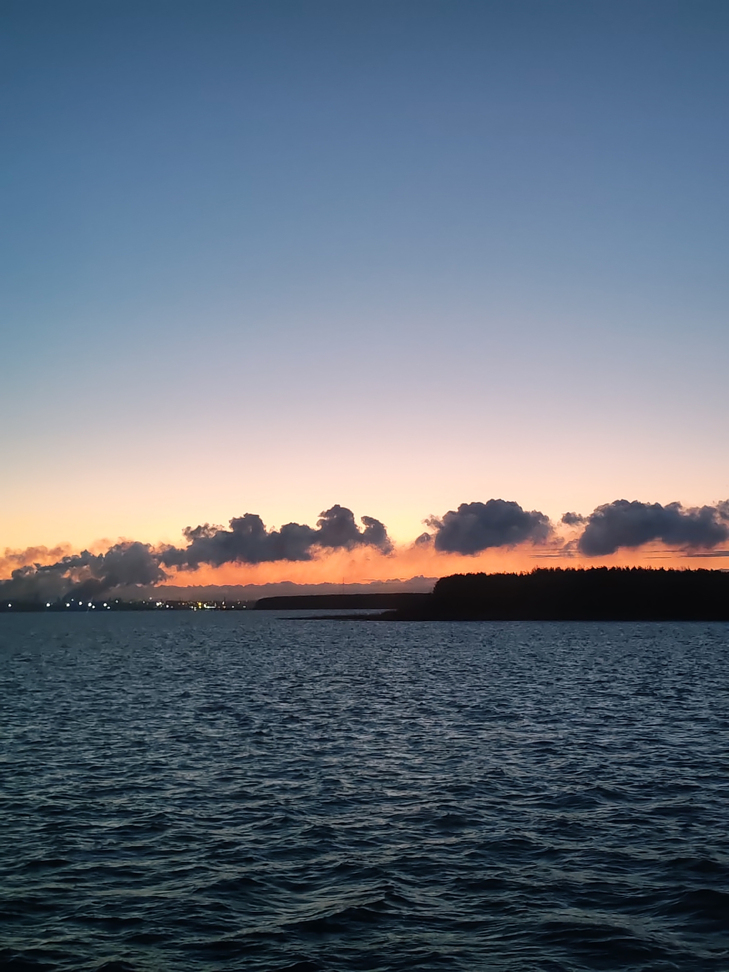 The image depicts a body of water with gentle ripples, likely a sea or a large lake, under a sunset or twilight sky. The sky transitions from a deep blue at the top to a soft orange-pink near the horizon. Along the horizon, there is a dark strip of land, possibly an island or the coastline, topped with a dense line of trees or forest. To the left of the horizon, there are some indistinct lights, potentially from a city or industrial area. Above these lights, there are scattered clouds that appear gray against the orange and pink sky, hinting at either pollution or natural cloud formations. The location is undetermined as the city and region are Unknown, Unknown.
