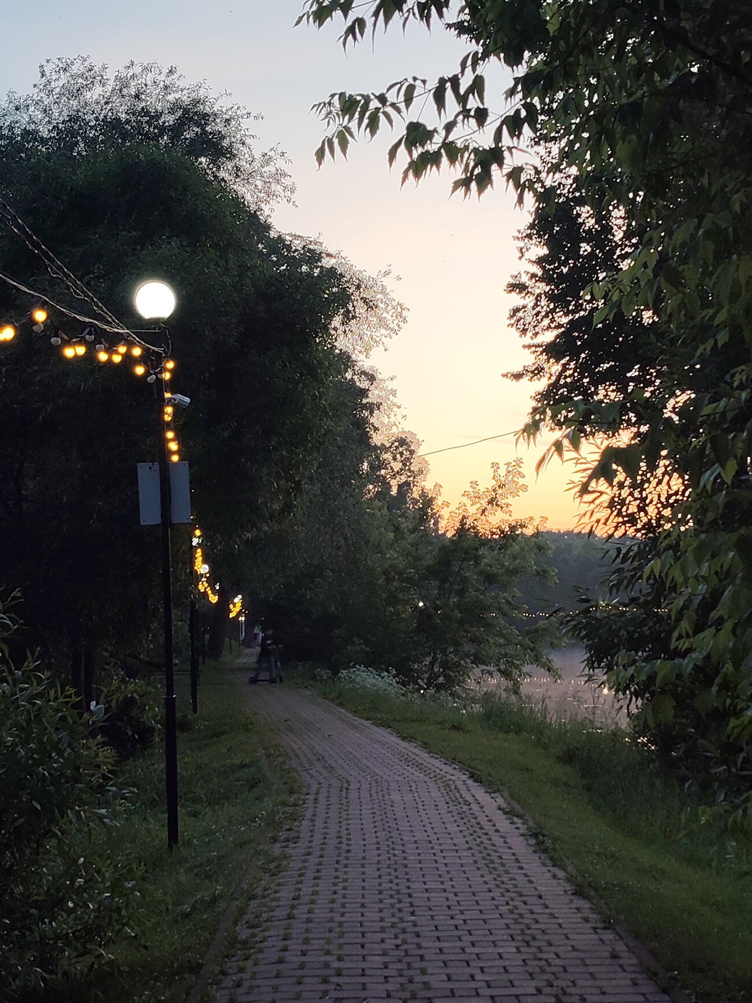 The image depicts a paved pathway in what appears to be a park or recreational area, possibly located in Chekhov, Russia, given the context. The pathway is constructed of interlocking bricks and is bordered by grass and dense foliage on either side. A lamppost with a spherical light fixture and a string of decorative lights illuminates the scene. Further down the path, a person on a scooter is visible. In the background, a body of water, likely a river or lake, can be seen, reflecting the soft hues of the twilight sky, which ranges from pale blue at the top to orange and yellow near the horizon. The overall atmosphere is tranquil, suggesting a late evening or early morning setting.