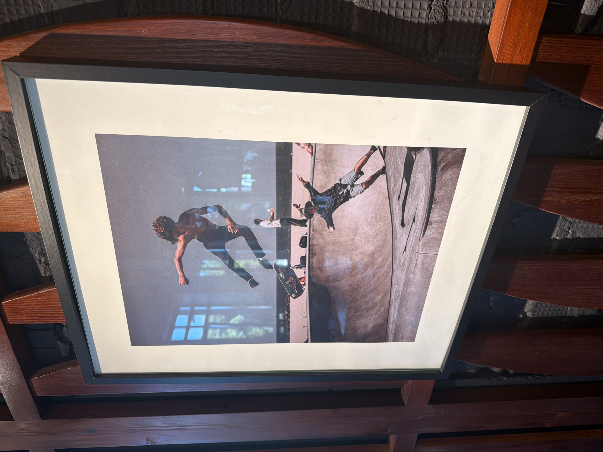 The image shows a framed photograph of a skateboarding event in a skatepark. The photograph is mounted on a wooden wall structure. In the foreground of the photograph, a skateboarder in a plaid shirt and shorts is captured mid-fall, with arms outstretched for balance. In the background, another skateboarder is airborne, performing a trick. The skatepark appears to be an indoor facility with a smooth, curved concrete surface. The background of the photograph shows large windows with a grid pattern, suggesting an urban setting. The wooden structure holding the photograph is made of dark red wood and is attached to a dark, textured wall. There are no visible elements in the image that indicate the location is in Kazan, Russia.