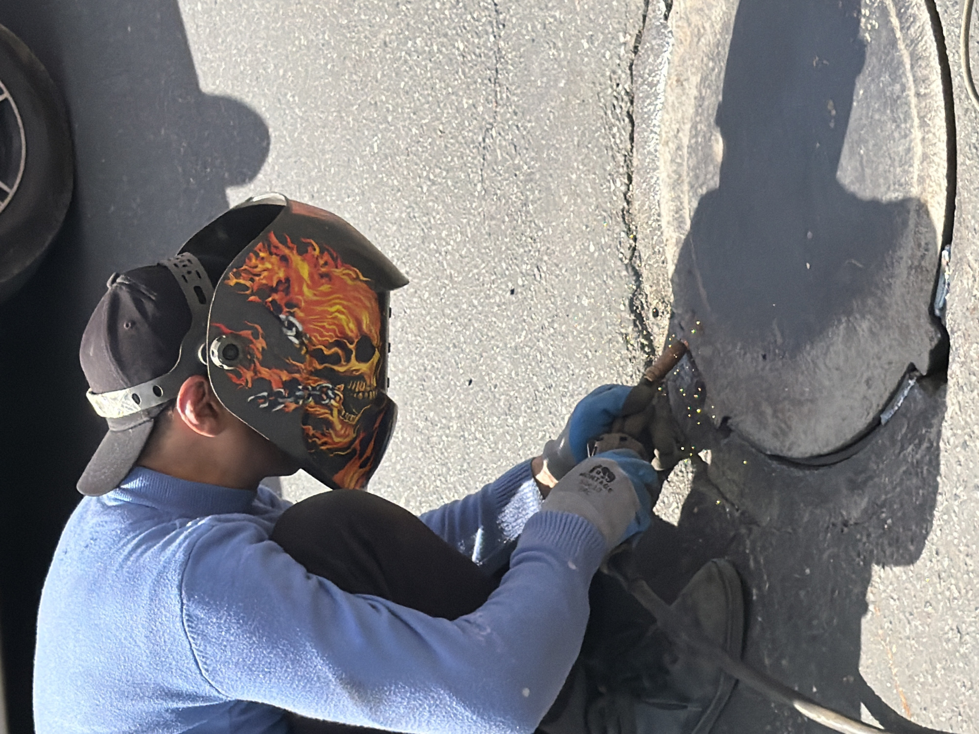 A person wearing a black baseball cap, a blue sweater, black pants, gray and blue work gloves, and a welding mask with a skull and flames design is welding a black metal object outdoors on asphalt. The mask obscures the person's face. Sparks are visible from the welding process. The scene appears to be set outdoors. The exact location is not discernible from the image.