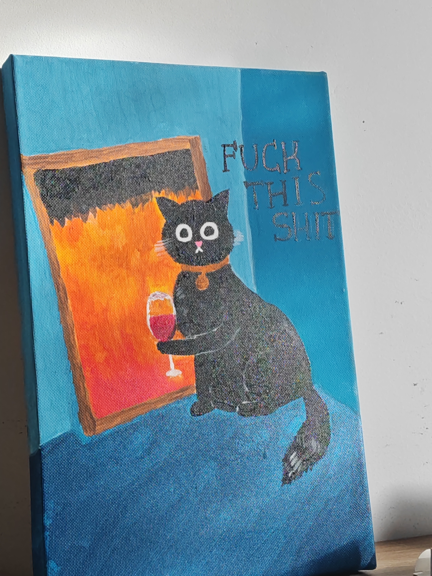 The image is a painting on a canvas featuring a black cat with large white eyes and a pink nose, wearing a collar. The cat is holding a glass of red wine in its right paw and is depicted sitting on a blue surface. To the cat's left is a rectangular frame with a fiery orange and black interior, possibly representing a fireplace or a window with a dramatic view. The background is a solid blue, and the words "FUCK THIS SHIT" are painted in silver letters in the upper right quadrant of the canvas. The painting is lit from the left, casting a shadow on the right side. There is no indication of any specific location in the city of Đà Nẵng, Vietnam.