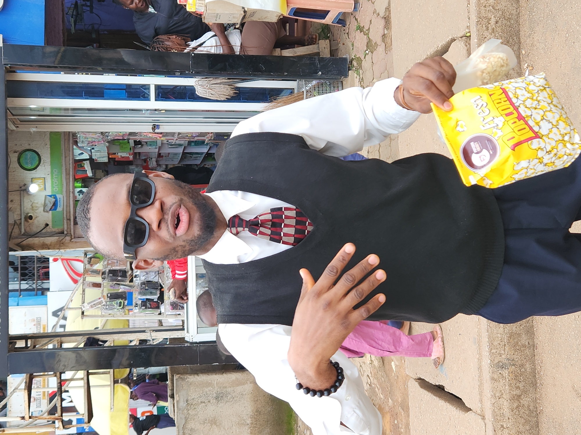 A man stands in front of a shop in Jos, Nigeria, holding a bag of popcorn. He wears sunglasses, a white shirt, a black vest, and dark blue pants. A woman with braided hair stands behind him in the shop entrance, and another woman sits in the background. The shop appears to sell mobile phone accessories, with items displayed on shelves and racks. The ground is paved with stones, and there is a concrete barrier in the foreground.