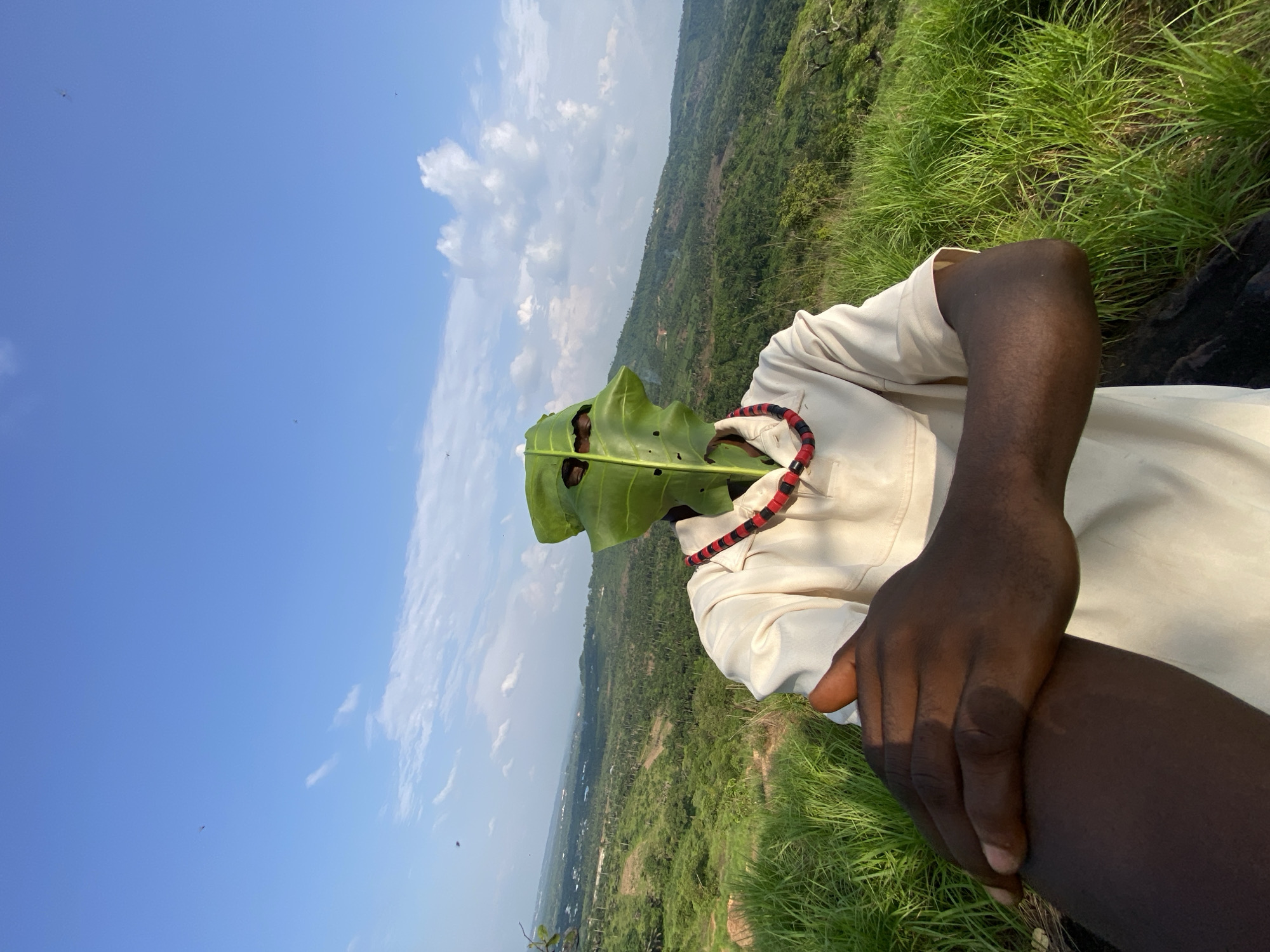 A person is standing on a grassy hill overlooking a valley, wearing a light-colored shirt and a necklace. A large green leaf is positioned in front of their face, obscuring it and creating eyeholes. The person's left arm is crossed over their chest, with their hand resting on their bicep. The background shows a lush green landscape with rolling hills and a blue sky with scattered clouds. The location is identified as Achara, Nigeria.
