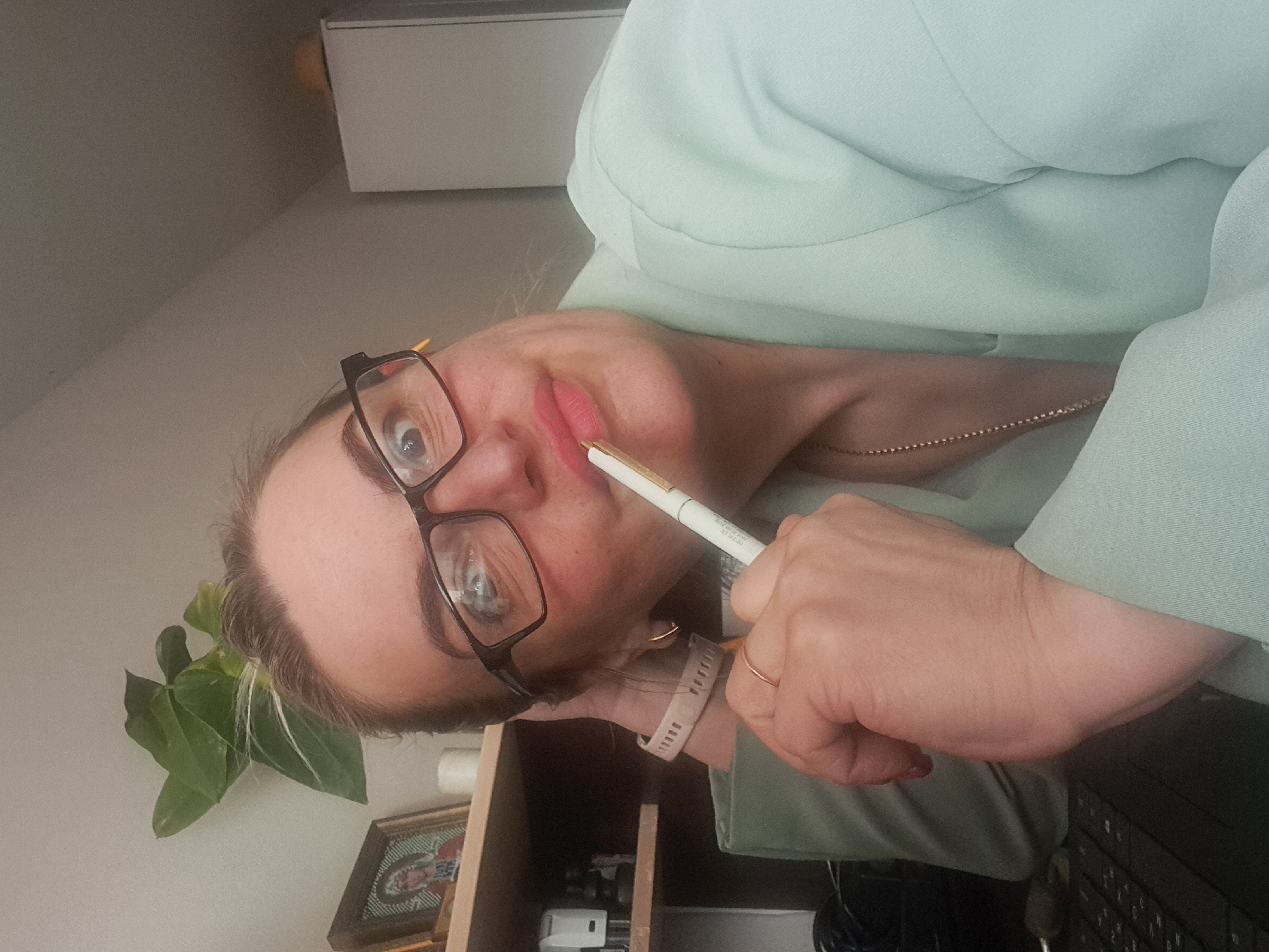 A woman with blond hair, wearing eyeglasses and a light green blazer, is holding a pen to her lips. Her right hand is visible, with a ring on the ring finger and a watch on the wrist. She appears to be indoors in a room with a desk and a plant. A picture frame is visible on the desk. A white box or cabinet is also partially visible. The image orientation is rotated, possibly taken with a phone. There is no information to conclude this is Elektrostal, Russia.