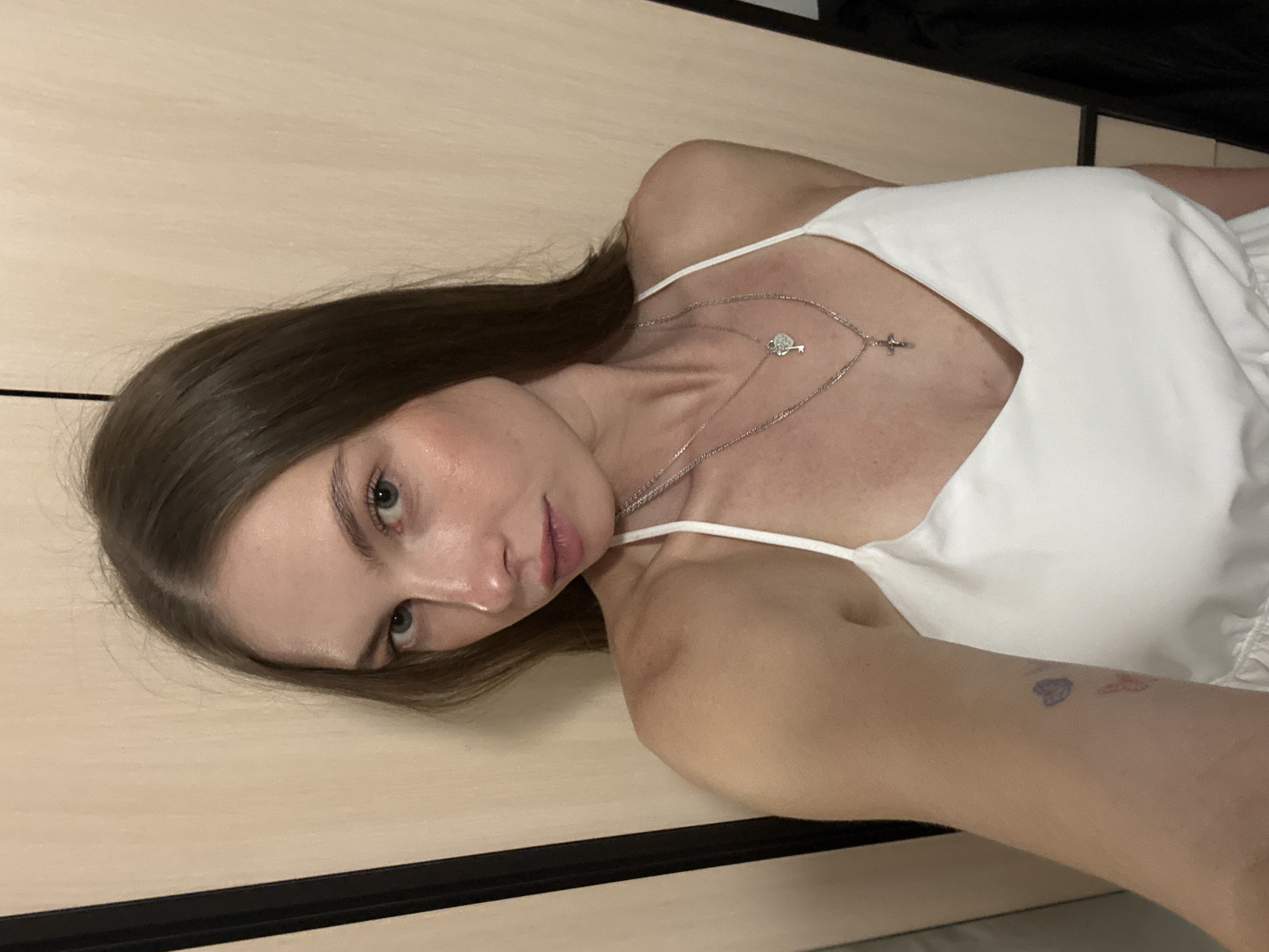 A young Caucasian woman with long, dark brown hair is posing for a selfie in a room with light-colored, wood-paneled walls. She is wearing a white, v-neck dress with spaghetti straps and two silver necklaces, one with a heart-shaped pendant and a key, and the other with a cross. There are small, colorful tattoos on her left arm, including a purple heart and an orange butterfly. The woman's facial expression is neutral. The background consists of two vertical, dark panels on the left and right edges of the frame, suggesting she is standing in a doorway or between two pieces of furniture. The image does not provide enough information to determine the specific location in the city of Perm, Russia.