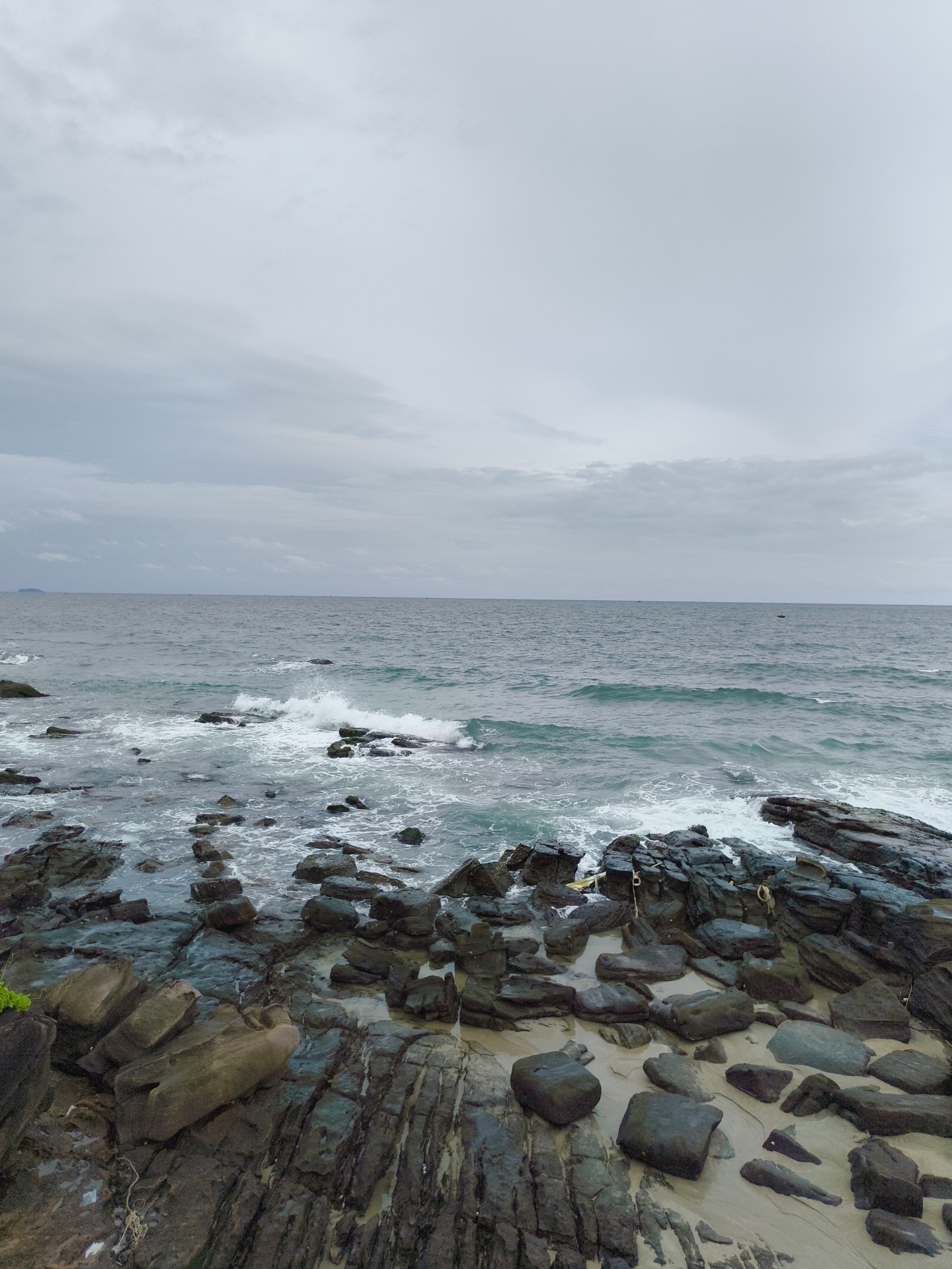 The image depicts a rocky shoreline on a cloudy day. The foreground is dominated by large, dark-colored rocks and stones scattered across a sandy beach, with some vegetation visible on the far left. The sea is a muted blue-green, with white waves crashing against the rocks. The horizon is visible, with a faint landmass in the distance on the left, and a small boat can be seen on the right. The sky is overcast with a pale gray hue. The overall impression is one of a natural, somewhat rugged coastal landscape. This scene is consistent with the rocky coastlines found in areas like Sao Beach on Phu Quoc Island, Vietnam, known for its natural beauty and varied geological features.