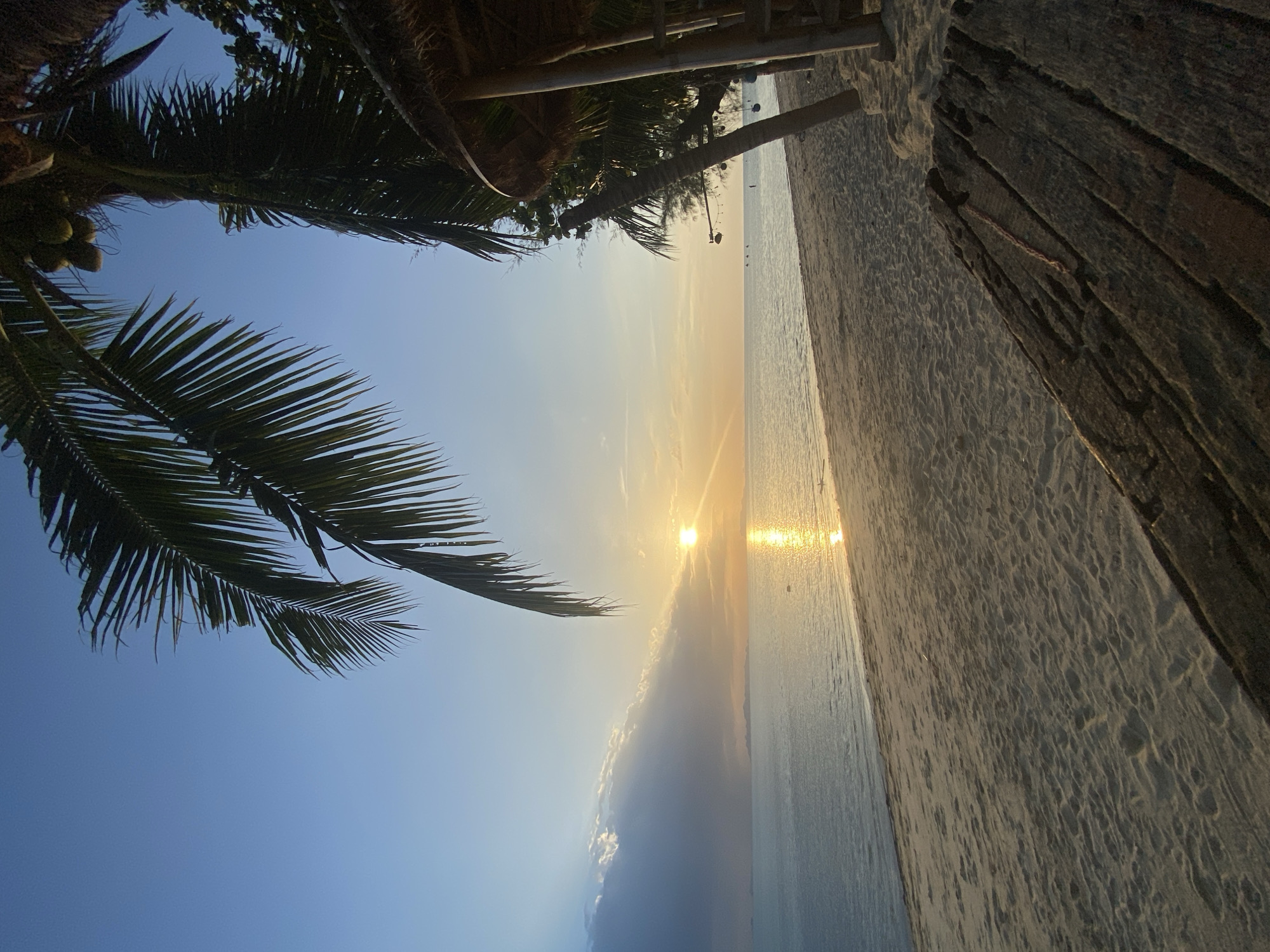 An outdoor, eye-level, vertical, full shot captures a serene beach scene at dusk on Ko Samui, Thailand. The setting sun casts a warm, golden glow across the calm ocean and the sandy shore. In the foreground, a weathered wooden structure, possibly part of a pier or seating area, occupies the lower right quadrant. The beach is composed of fine, light-colored sand with visible footprints. Lush palm trees, their fronds silhouetted against the sky, frame the upper left and right portions of the image. To the left, a section of clouds is illuminated by the sun's rays, creating a soft, diffused light. In the distance, faint silhouettes of islands or landmasses can be seen on the horizon. The ocean is a deep blue, reflecting the golden hues of the sunset. A small thatched-roof structure is visible on the right side of the frame, suggesting a beachside establishment. The overall atmosphere is peaceful and tranquil, indicative of a tropical evening.