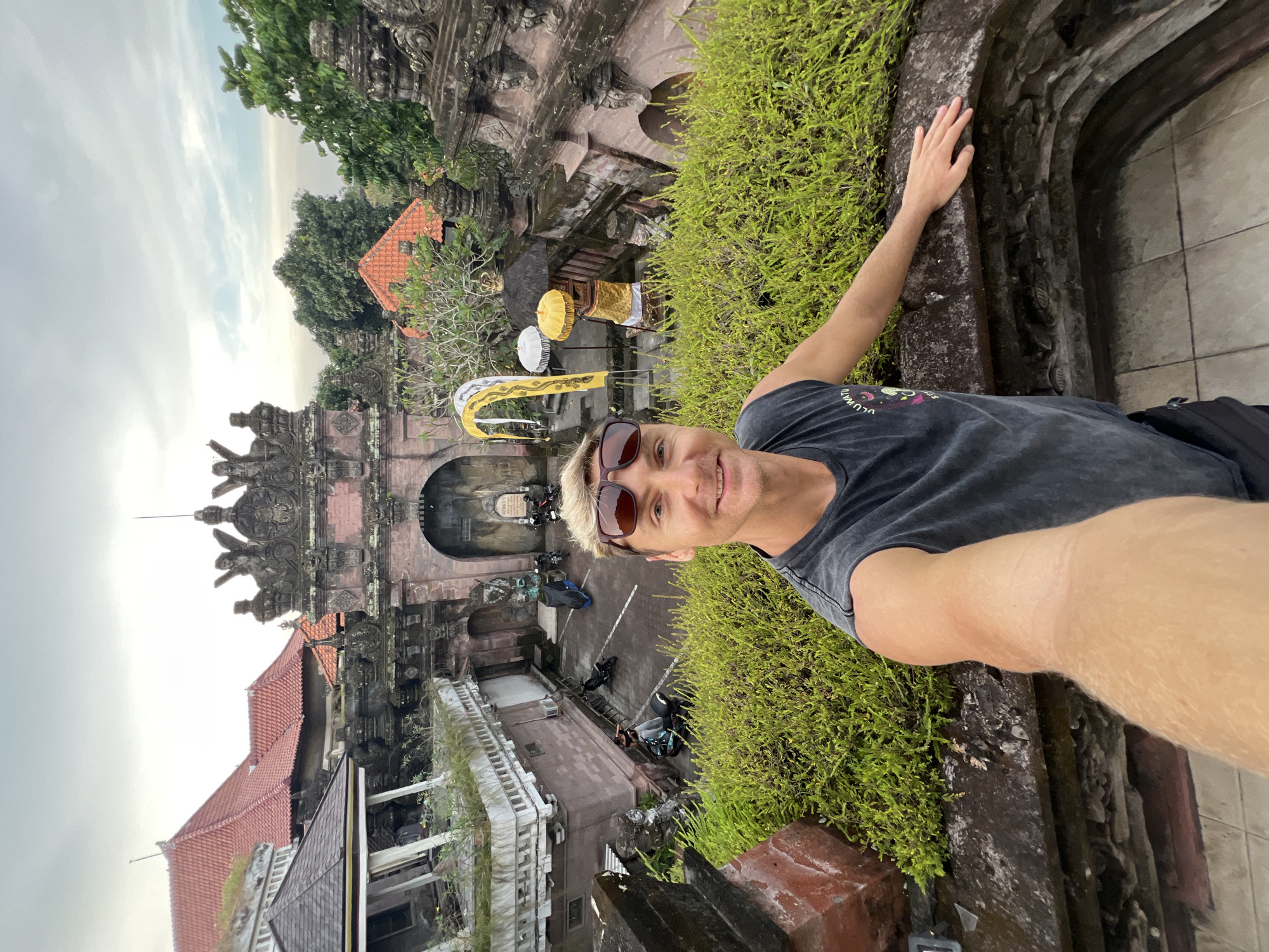 A man is taking a selfie at a location in Celuk, Indonesia. He is wearing sunglasses and a tank top and standing near a stone wall covered in green plants. In the background is a traditional Balinese gate with intricate carvings, red tiled roofs, and decorative umbrellas. There are motorcycles parked in the area behind the gate. The sky is overcast.