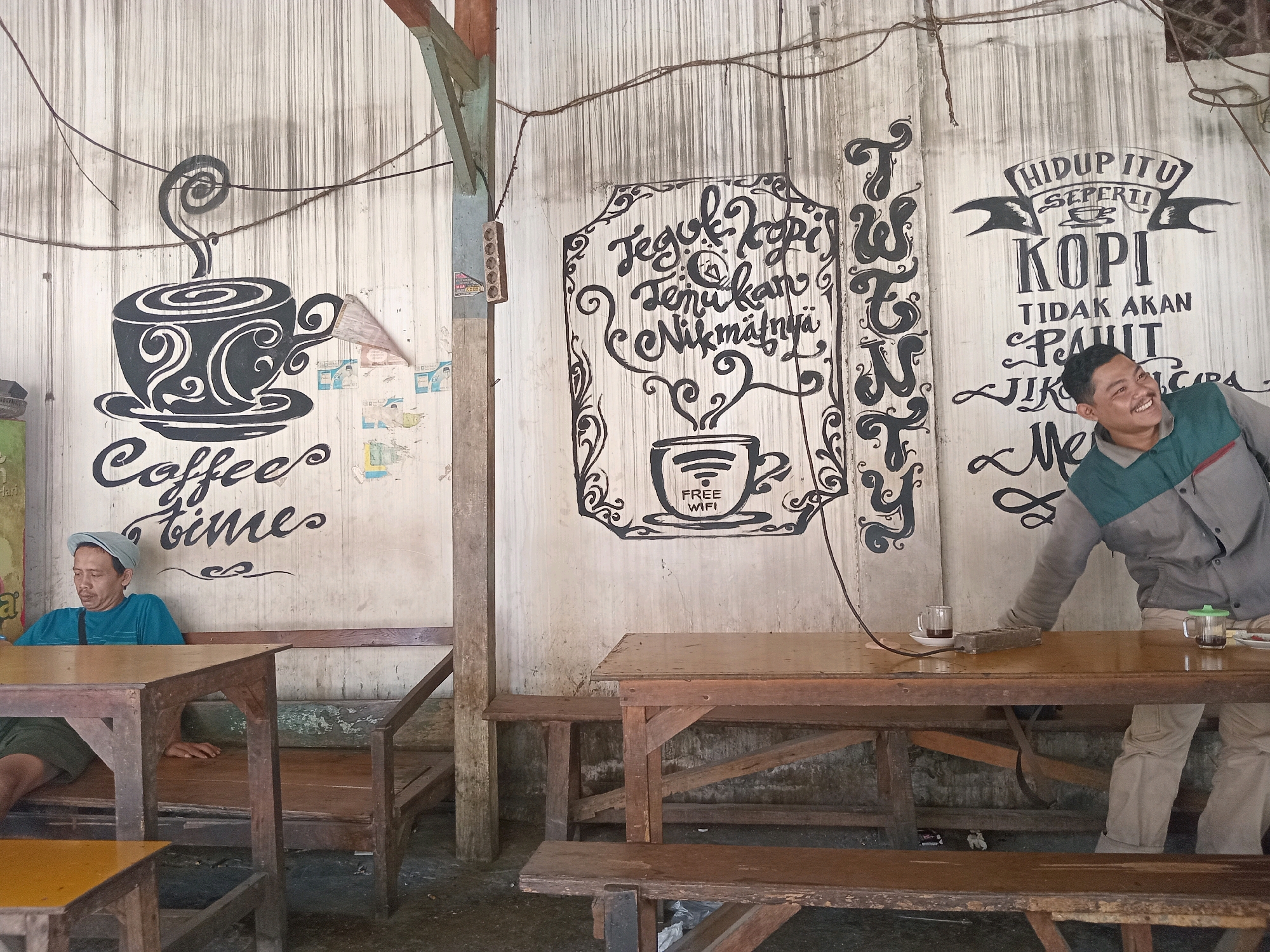 The image depicts a rustic outdoor cafe in Ngipik, Indonesia. On the left, a man wearing a blue t-shirt and a light-colored cap sits at a wooden table and bench, facing left. To the right, another man, dressed in a grey and green jacket and light-colored pants, leans over a wooden table, seemingly wiping it with a cloth. He is smiling and looking to his right.
The cafe's wall is adorned with hand-painted signage. On the left, there's a stylized drawing of a coffee cup with the words "Coffee times" below it. In the center, a decorative frame surrounds text that appears to be in Indonesian. To the right, a banner reads "HIDUP ITU SEPERTI KOPI" (Life is like coffee), with further text below. Two glasses of coffee are visible on the tables, one with a green lid.
The environment suggests a casual, perhaps locally run establishment, with wooden furniture and a weathered, textured wall. Overhead, electrical wires are visible. The ground appears to be concrete or packed dirt.