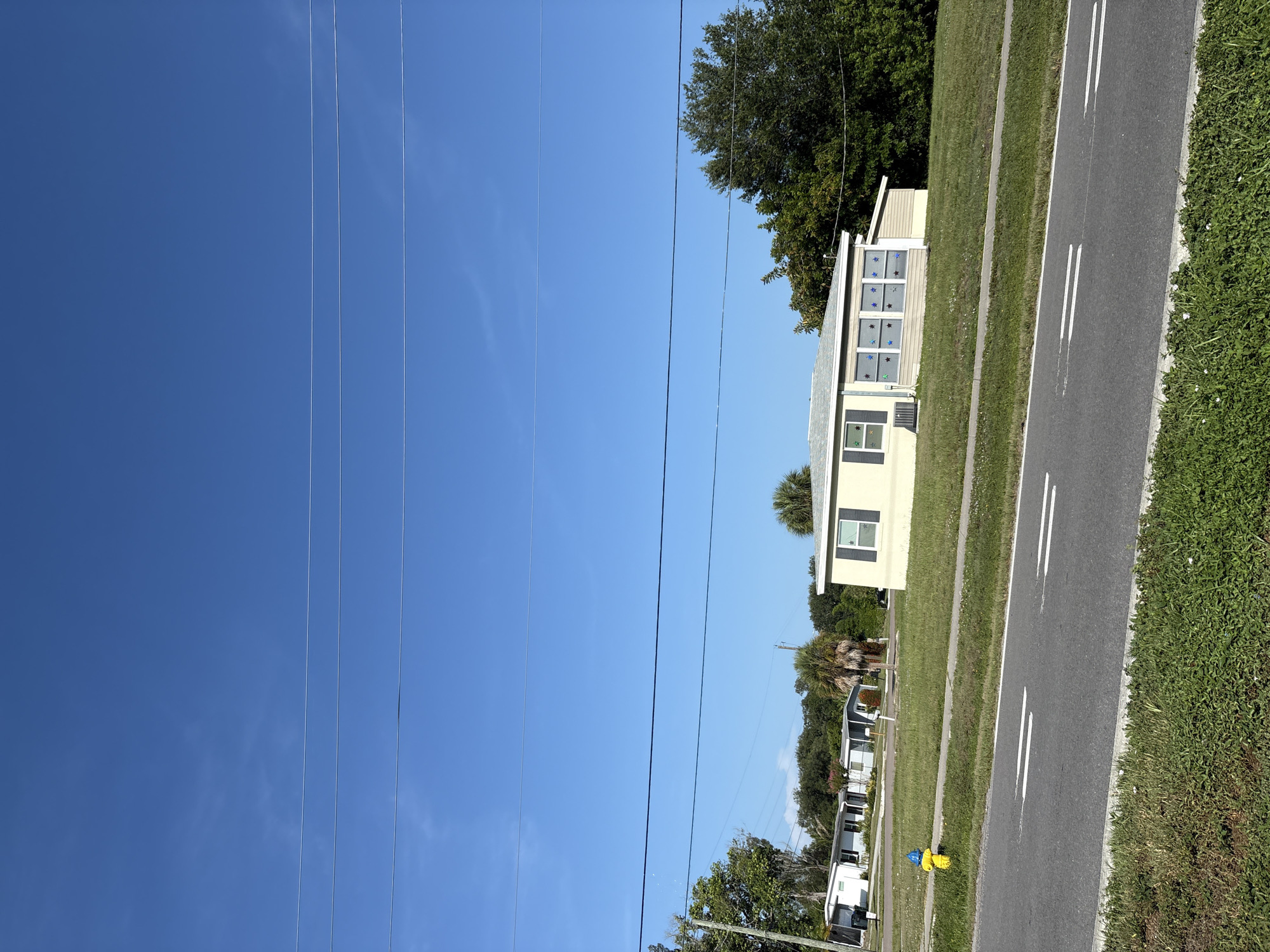 This is a daytime, eye-level, exterior, medium shot of a residential area on a clear day. In the foreground, a two-lane asphalt road runs horizontally across the frame, with a white double line painted in the center of each lane. The road is bordered by a strip of green grass. Beyond the road, a manicured lawn slopes upward toward a single-story tan house with a light gray roof. The house has a blue shutter on the left side of its front facade and a section with multiple windows decorated with blue star-shaped cutouts. An air conditioning unit is visible below the blue shutter. To the left of the house and closer to the road, a yellow and blue fire hydrant stands near the edge of the lawn. In the background, several other modest single-story houses are visible, some with white siding and others with light blue siding, interspersed with palm trees and other green foliage. Overhead, three thin, dark power lines stretch horizontally across the bright blue sky, with no clouds visible. The image appears to be taken in North Port, Florida, United States of America.
