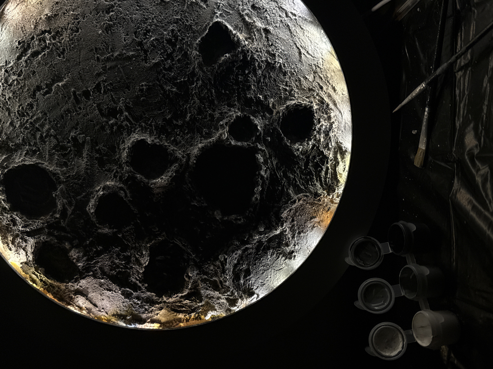The image depicts a model of the moon resting on a dark surface. The model is illuminated from behind, highlighting its cratered surface. To the right of the moon model are painting supplies, including paintbrushes and small containers of paint. The scene appears to be a workspace or studio. There is no readily available information to confirm that this scene is located in the city of Chaikovsky, Russia.