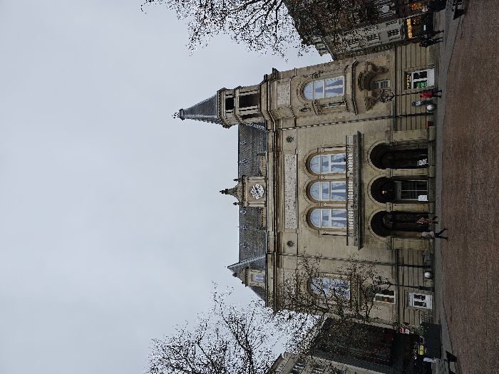 An external, eye-level photograph depicts the Cercle Cité building, located on Place d'Armes in Luxembourg City, Luxembourg, under an overcast sky. The prominent, multi-story structure is constructed from light-colored stone, featuring a steep dark roof with dormers, and a clock face centered on its main facade below a decorative pediment. To the right, a multi-tiered tower with a pointed spire rises. The building's ground level presents three large arched entrances, topped by a balcony with a stone railing. Above the balcony, three large arched windows are visible, and additional windows are present across the facade and a rounded bay on the right side.
In the foreground, a reddish-brown paved square extends across the frame. Several individuals are visible on the square: two figures walk towards the building's central arches, one figure walks left to right near the lower left, and two indistinct figures are near the building's right side. Bare deciduous tree branches partially obscure the upper left and right corners of the image. Surrounding the main building are other multi-story city buildings, partially visible on the left and right sides of the frame. Street furniture, including dark benches and a street lamp, is present on the square. The overall scene indicates an active urban environment on a dull, cloudy day.