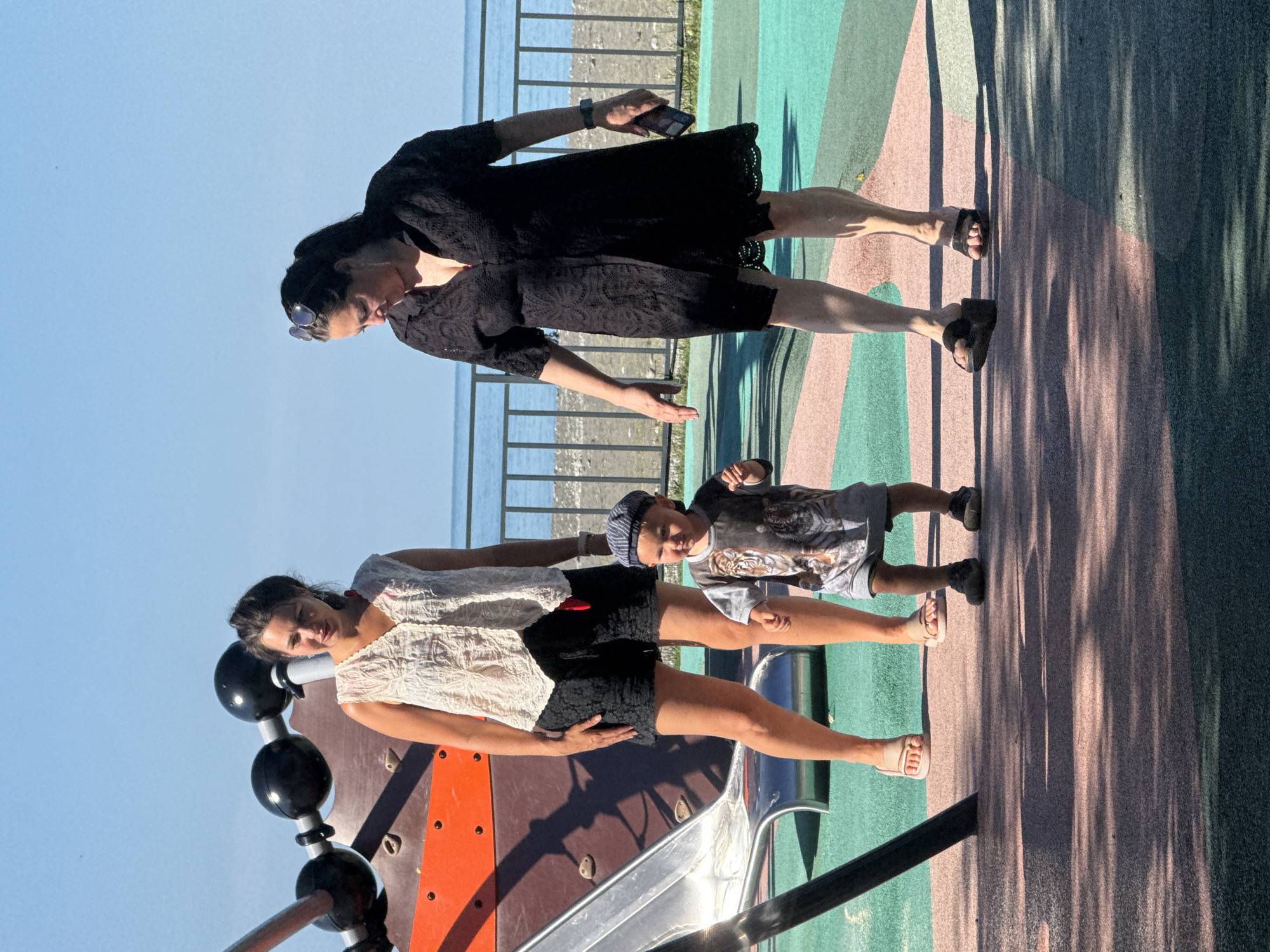 In Sochi, Russia, on a bright sunny day, two women and a young child are on a playground with a black fence in the background, separating the playground from a beach and the sea. The playground surface is a multi-colored mat, with a slide and part of a climbing structure visible on the left. The woman on the left is wearing a white sleeveless top and black shorts, with her dark hair pulled back. The woman on the right is wearing a black lace dress and sunglasses perched on her head. The child, positioned between the two women, is wearing a grey t-shirt with a tiger print and a blue and white striped cap, and is looking to the right. Both women appear to be standing still, while the child is mid-stride, as if walking or running. The woman on the right is holding a dark-colored smartphone in her right hand.