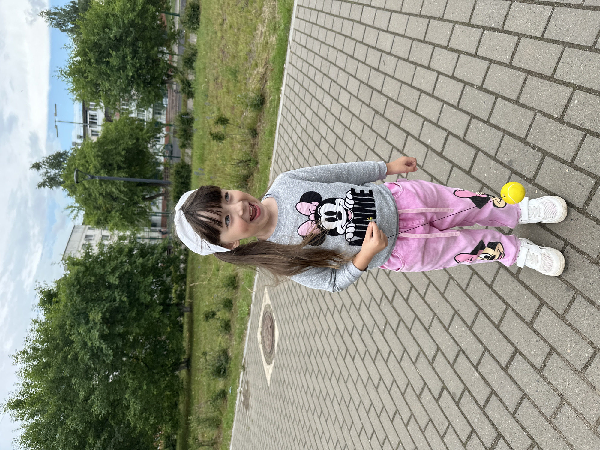 A young girl with dark hair pulled into two ponytails and wearing a gray sweatshirt with a Minnie Mouse graphic and pink pants with Minnie Mouse accents is standing on a paved walkway. She is holding a string with a yellow ball attached to it. The girl is smiling with her mouth open, looking to her left. In the background, there are trees, grass, a lamppost, and apartment buildings. The scene appears to be in a park or public outdoor space in the city of Chaikovsky, Russia. The sky is cloudy.